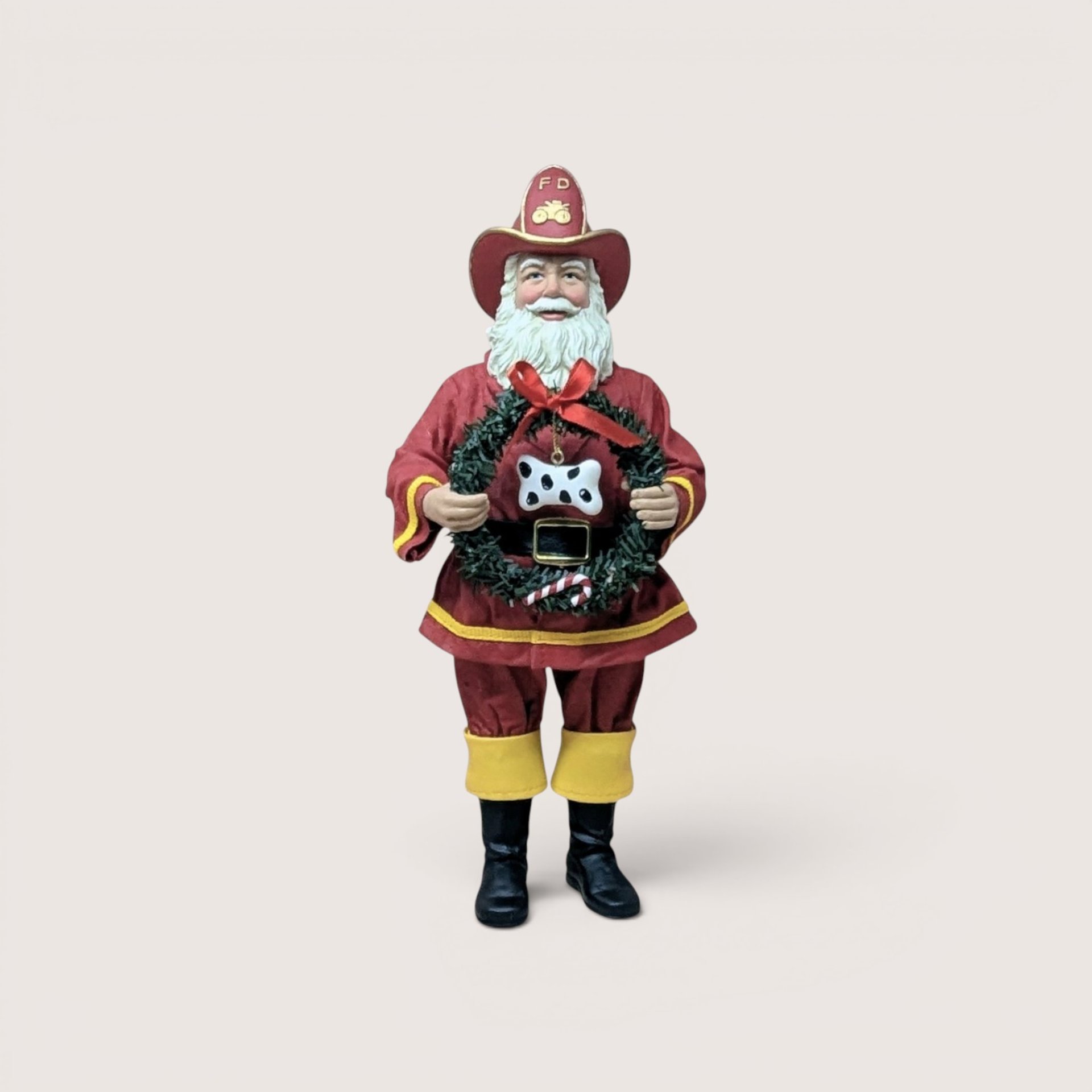 Vintage standing Santa Claus figurine dressed as a firefighter in a red turnout coat with yellow trim, black boots, and a matching red "FD" firefighter hat, holding a green Christmas wreath adorned with a red bow, a candy cane, and a black-and-white spotted dog bone ornament, centered on a plain linen background.