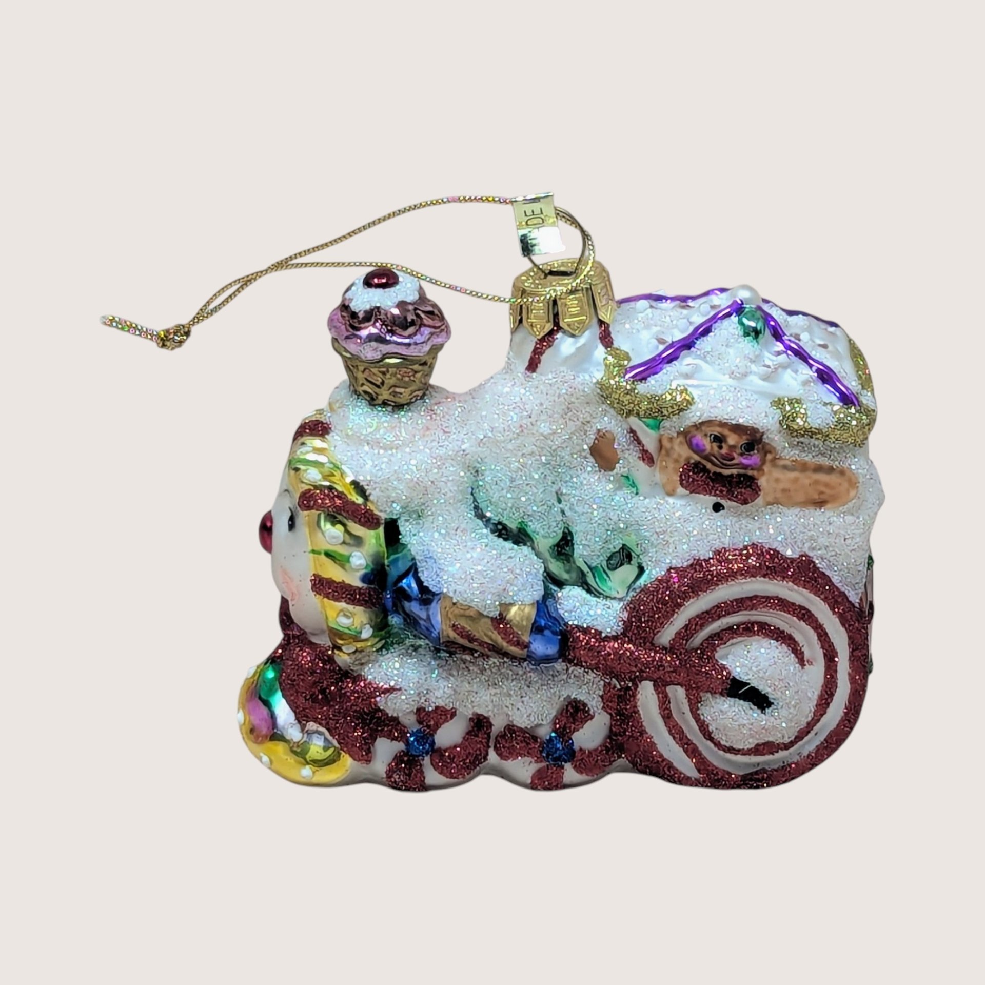 Vintage blown glass Christmas ornament shaped like a whimsical gingerbread or candy-themed locomotive train, heavily accented with white glitter snow, peppermint swirl wheels, and a cupcake smokestack, centered on a plain linen background.