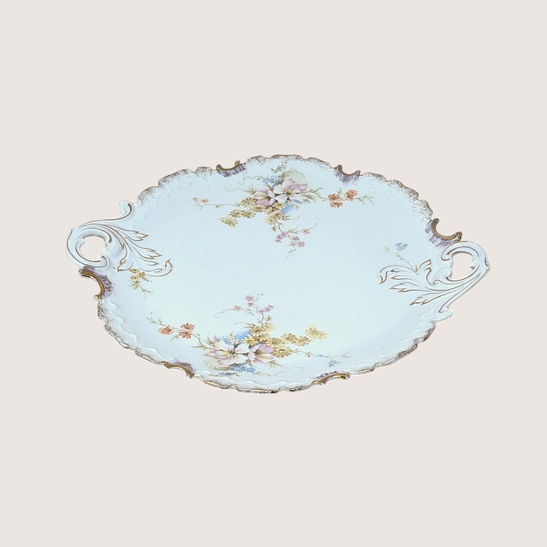 A vintage Rosenthal porcelain handled cake plate resting on a soft pink background. The antique German serving tray features delicate pink and yellow floral bouquets, an ornate scalloped edge, and brushed gold accents on the handles.