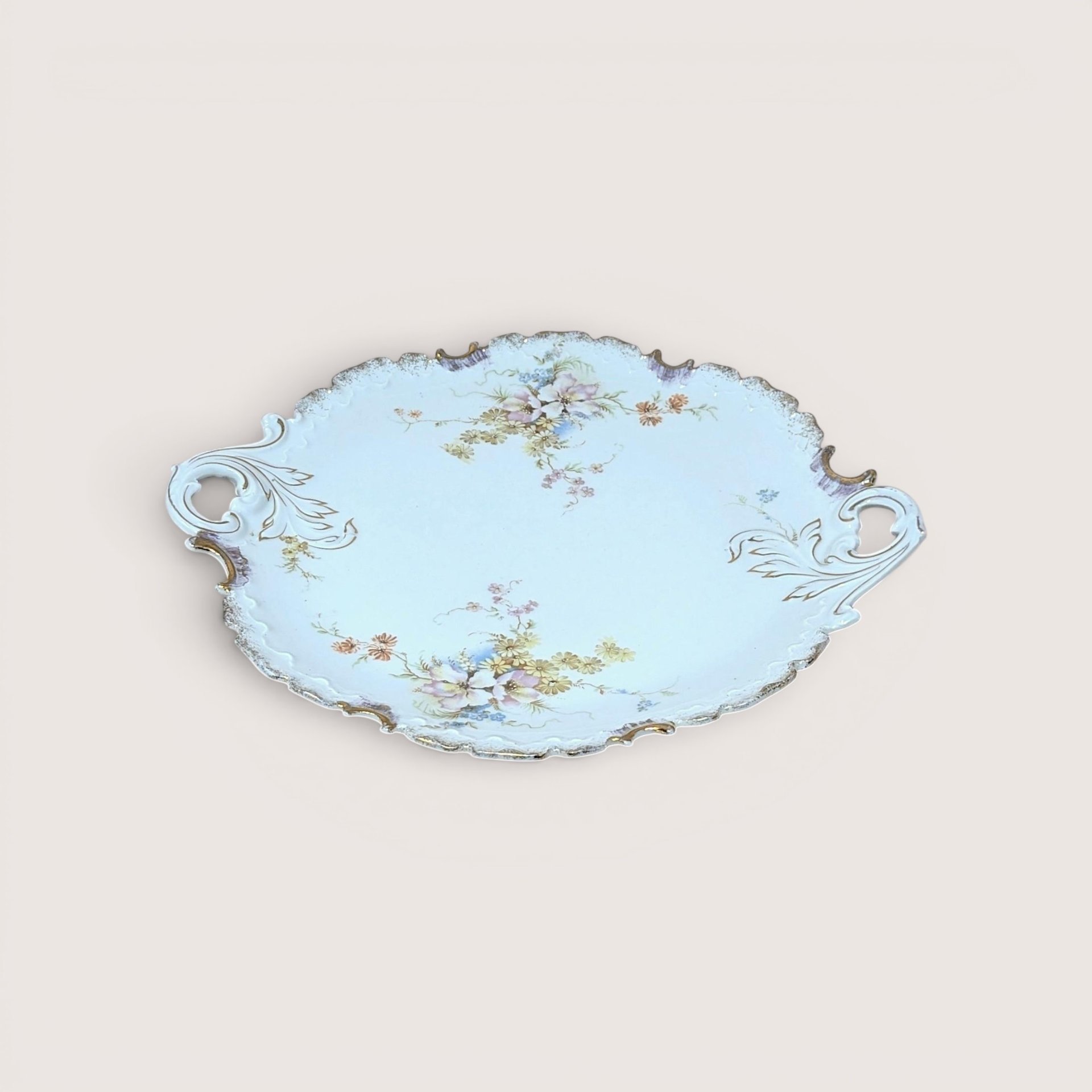 Vintage porcelain serving platter or cake plate with an elegant ruffled rim and twin integrated scroll handles, decorated with delicate hand-painted pastel floral sprays on a pale blue background with gold-gilt highlights, centered on a plain linen background.