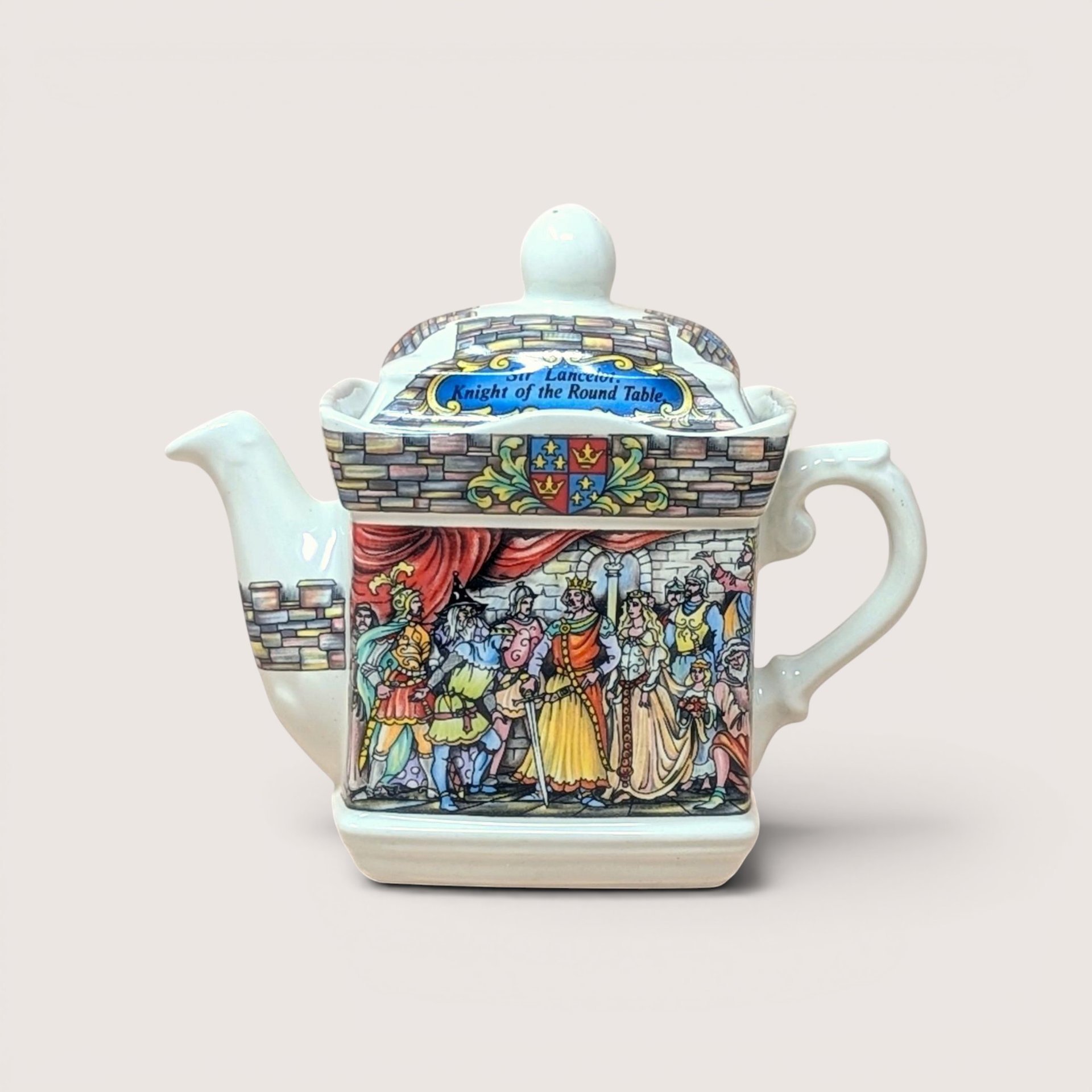 A vintage Sadler porcelain castle themed teapot on a soft pink background. The square cream colored teapot features a detailed double sided illustration of King Arthur's court at Camelot and Sir Lancelot with stone wall borders and a matching lid.