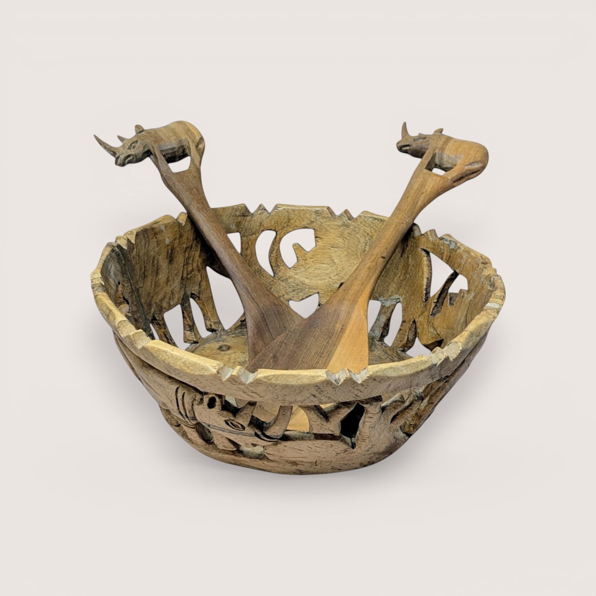 A vintage hand carved wooden safari bowl and matching serving utensils on a soft pink background. The openwork bowl features cutout silhouettes of elephants, while the two wooden servers have handles shaped like small rhinos.