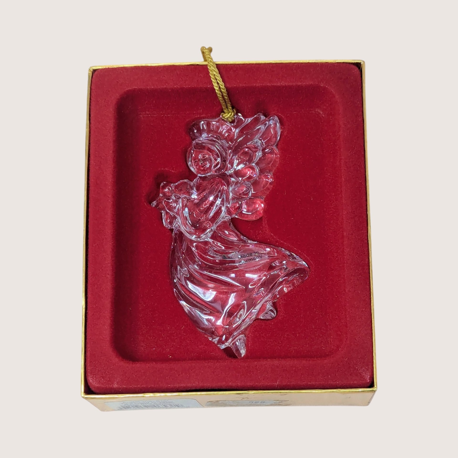 A vintage Gorham crystal angel ornament with a gold tassel on a soft pink background. The fine German glass sculpture features a clear, faceted angel in flight holding a small star, catching the light with brilliant clarity.