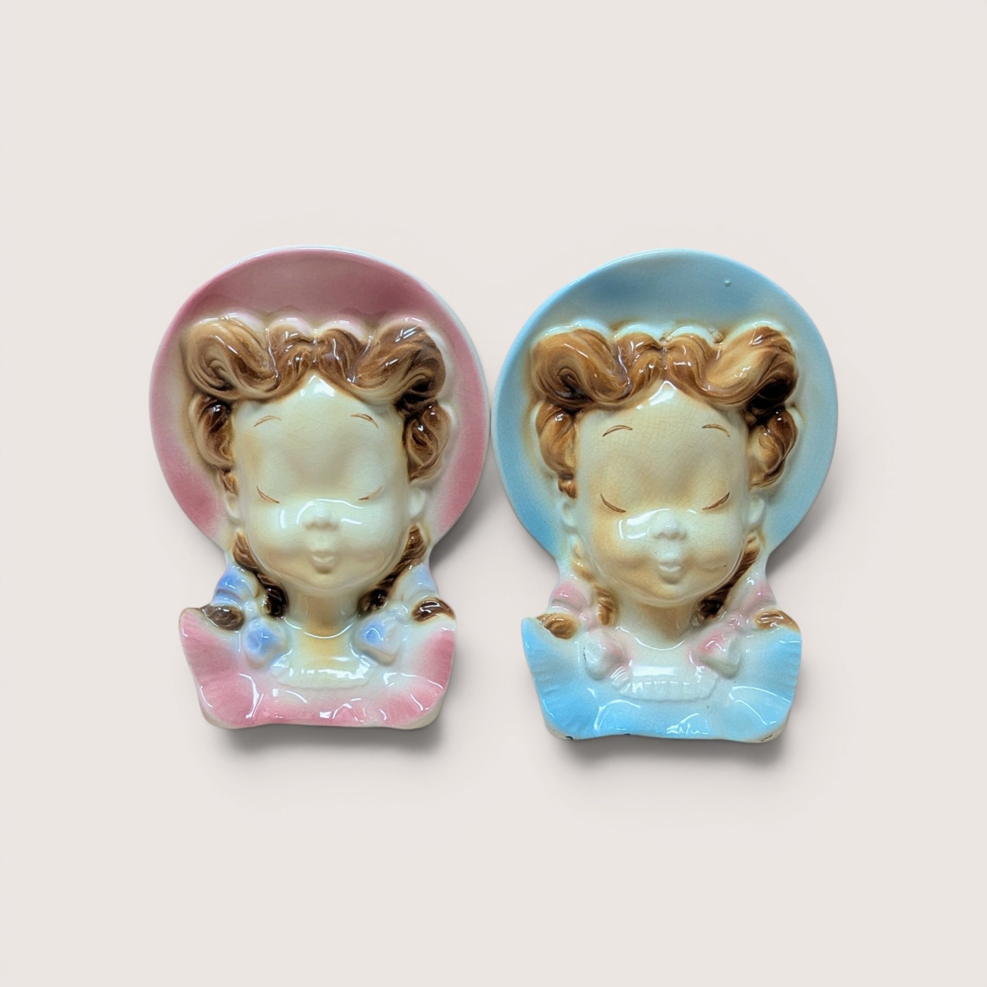Two vintage ceramic lady head wall pocket planters wearing pink and blue bonnets side by side on a plain background