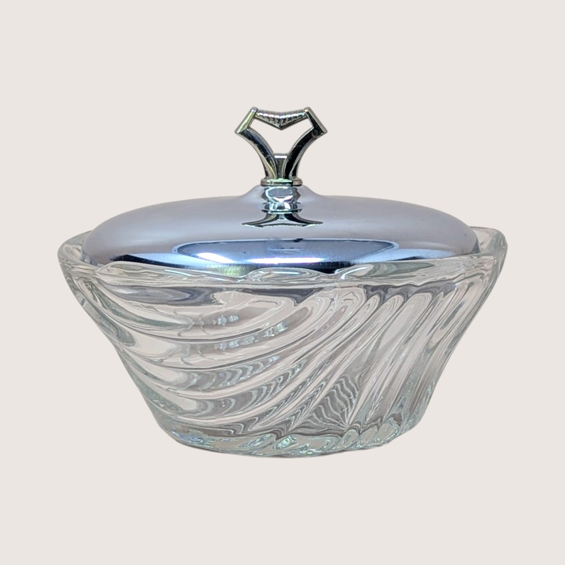 A vintage clear glass vanity bowl with a swirled pattern and a polished chrome lid featuring a geometric chevron handle.
