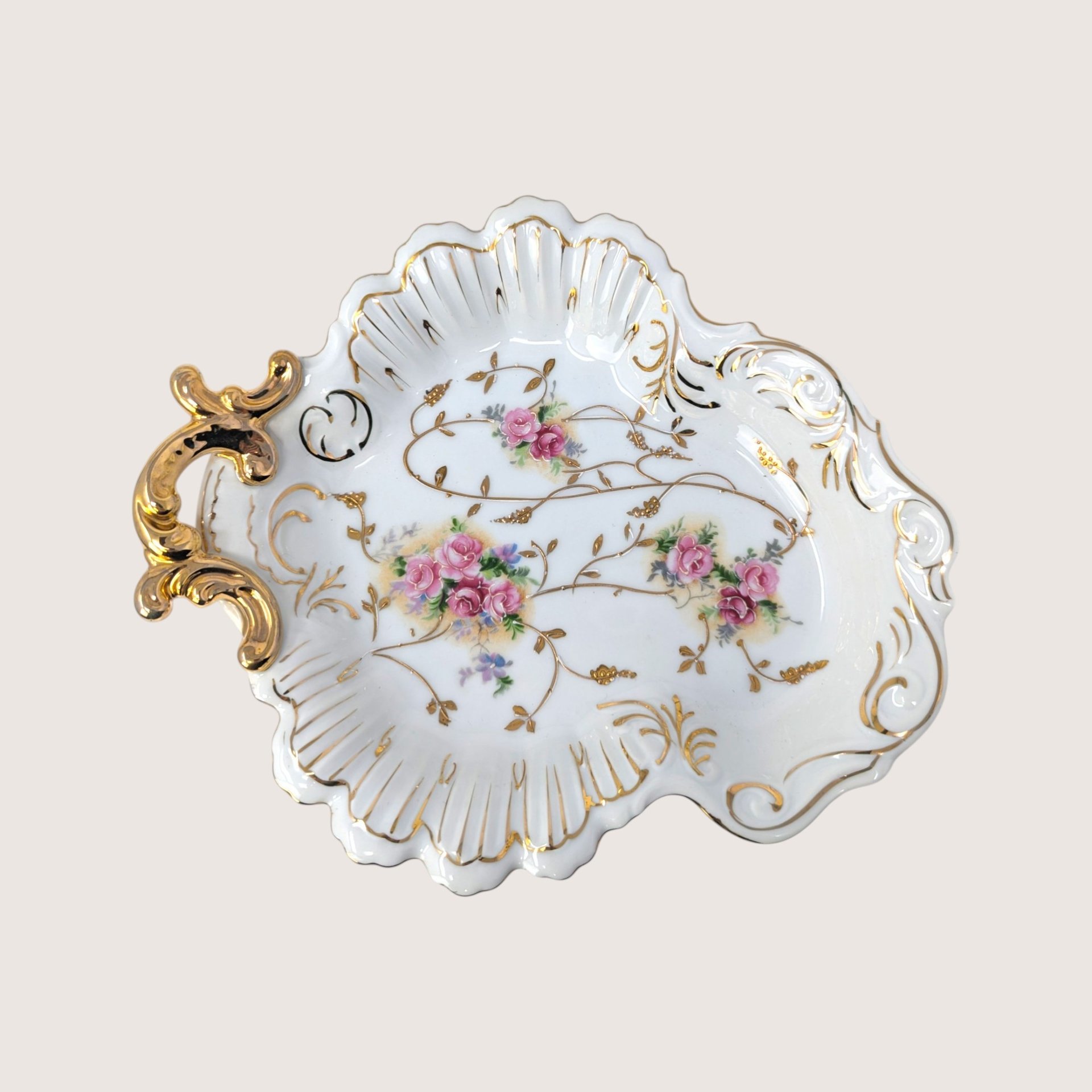 A vintage white porcelain shell dish with pink rose bouquets and an ornate gold handle on a plain cream background