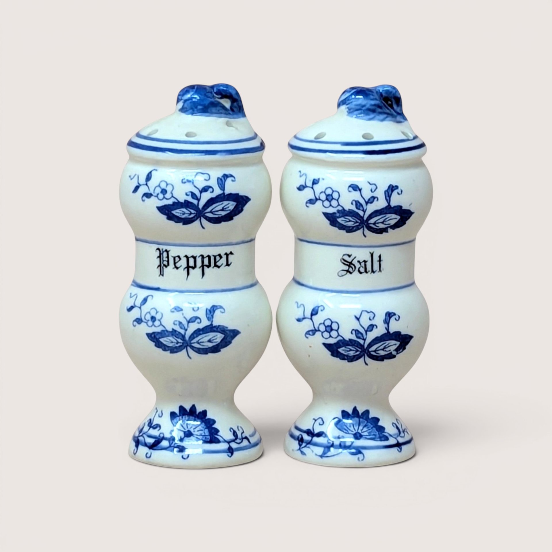 A pair of white ceramic hourglass shaped salt and pepper shakers with a blue onion pattern on a light beige backdrop