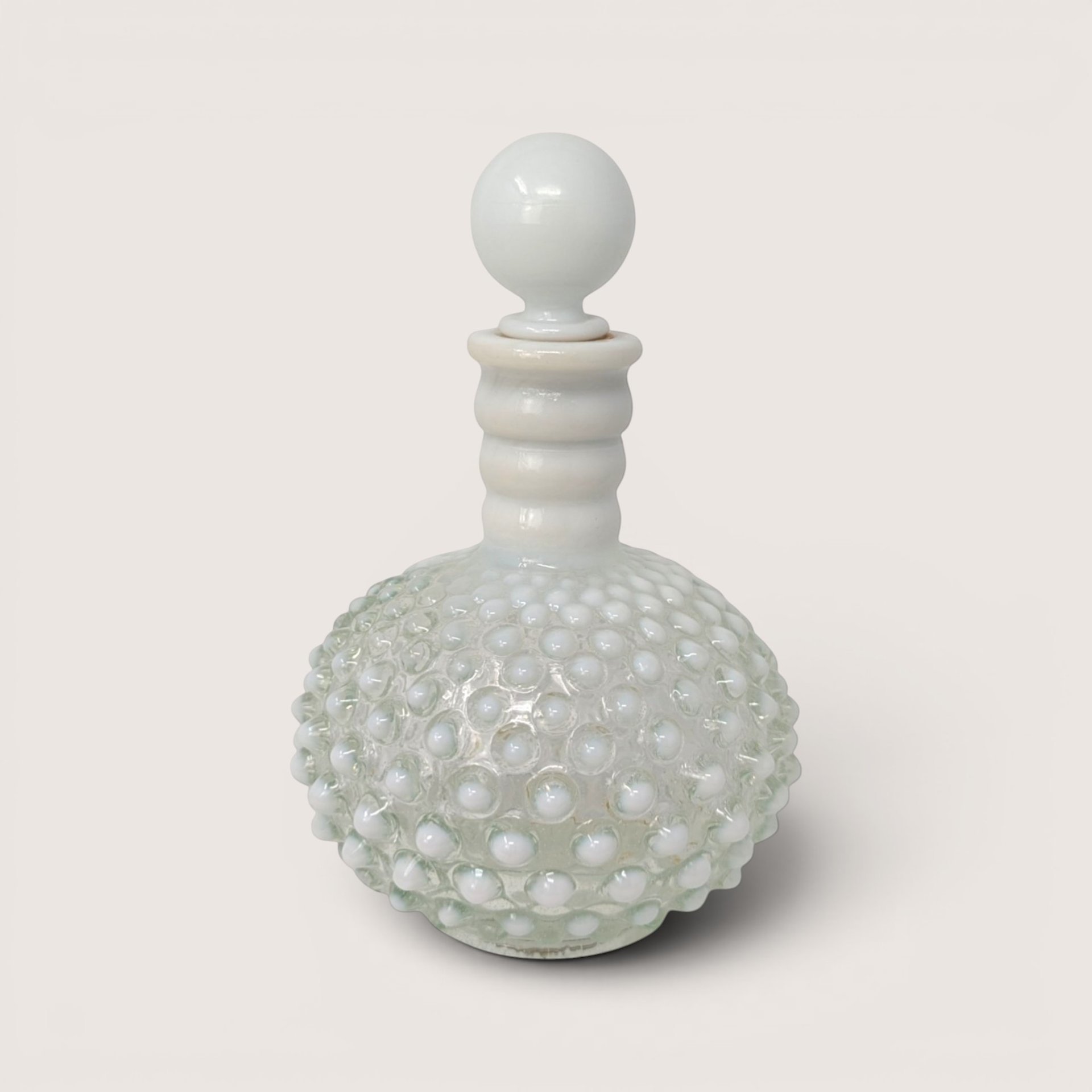 White opalescent hobnail glass cologne bottle with matching round stopper against a plain cream backdrop.