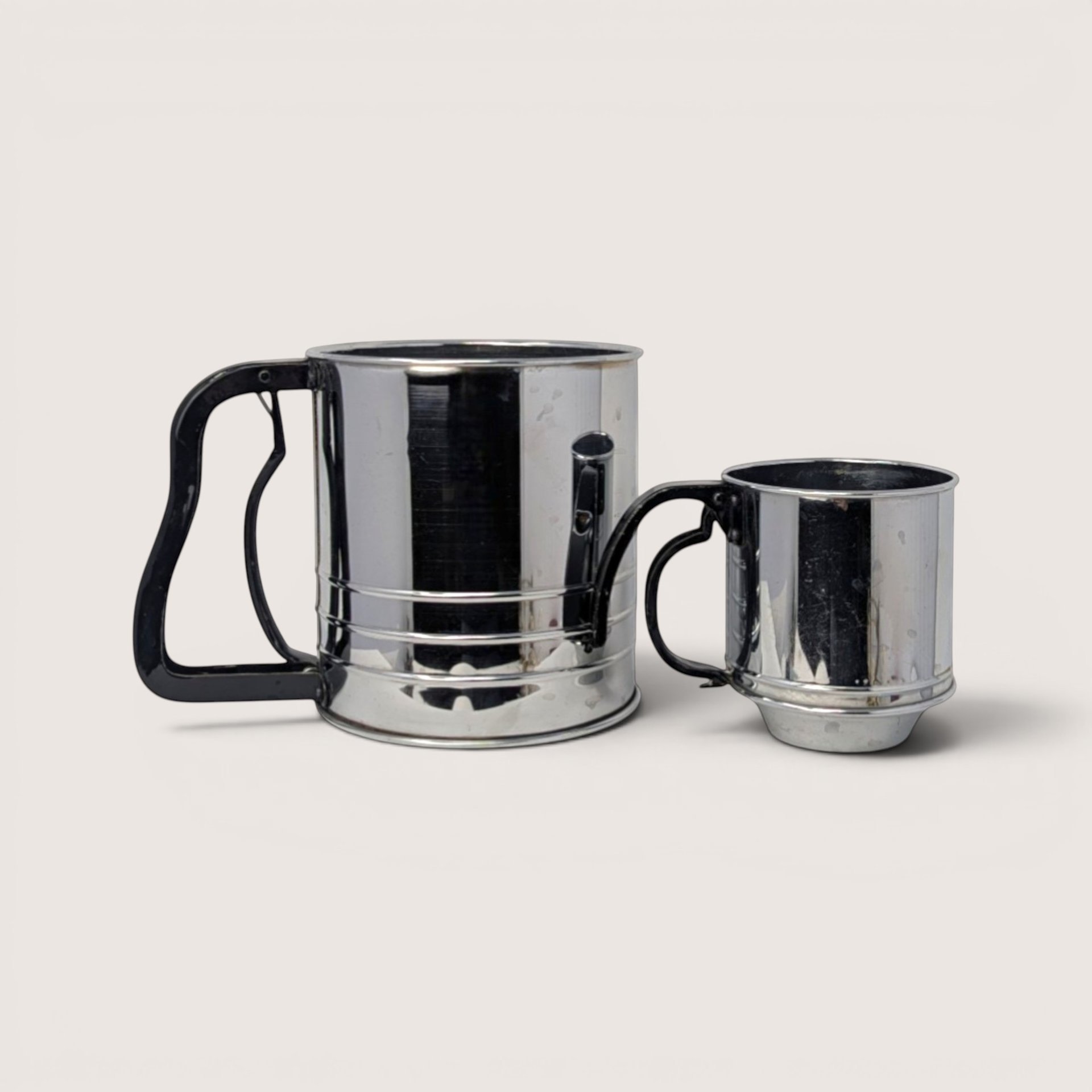 Two vintage stainless steel flour sifters with black squeeze handles side by side on a linen background.