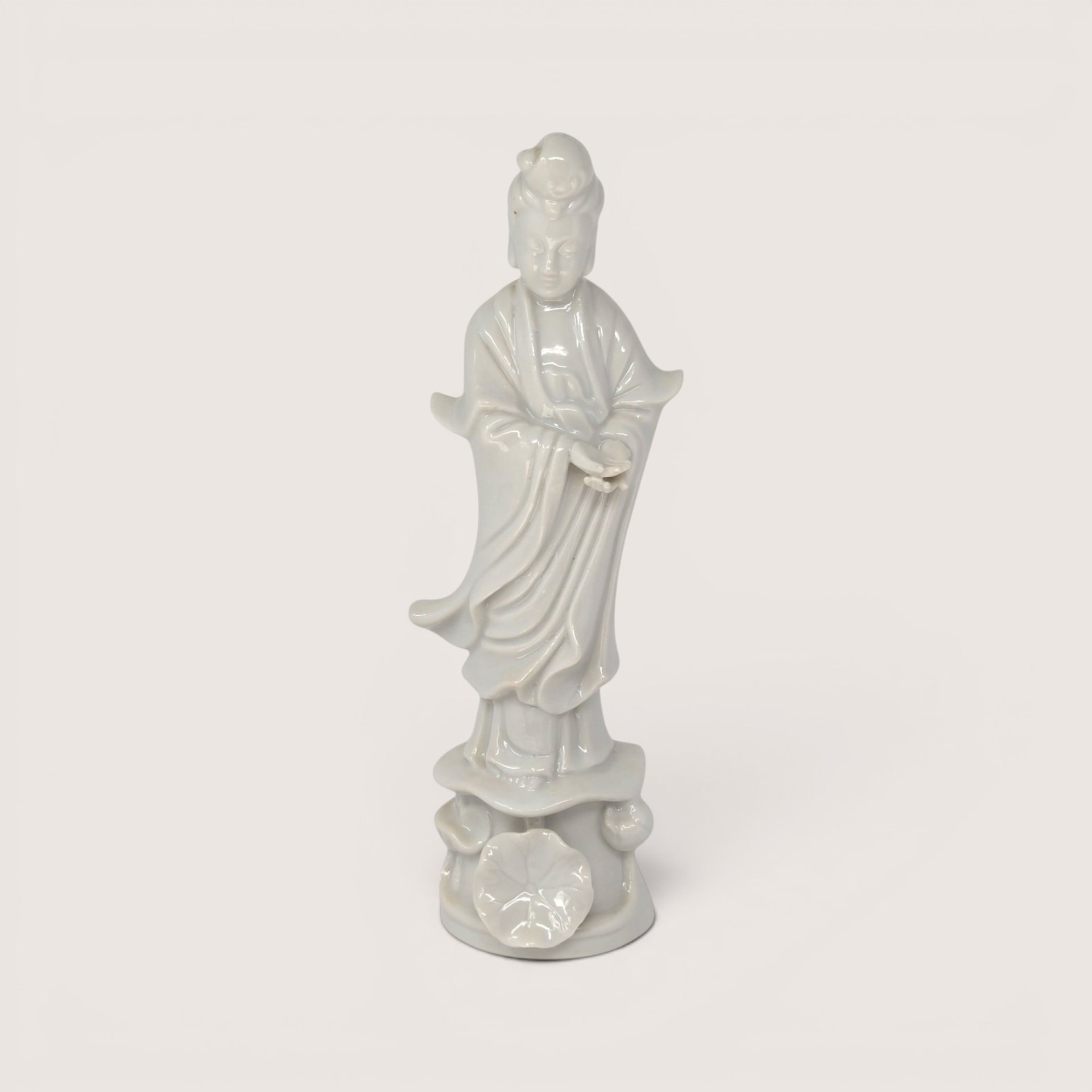 Vintage white porcelain Guan Yin statue standing on a sculpted lotus leaf pedestal base against a light background.