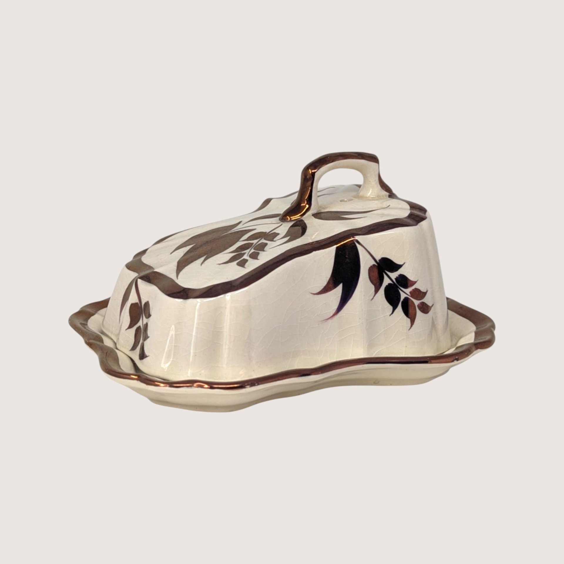 A vintage cream-colored ceramic butter dish with metallic copper luster leaf patterns and trimmed wavy edges.  