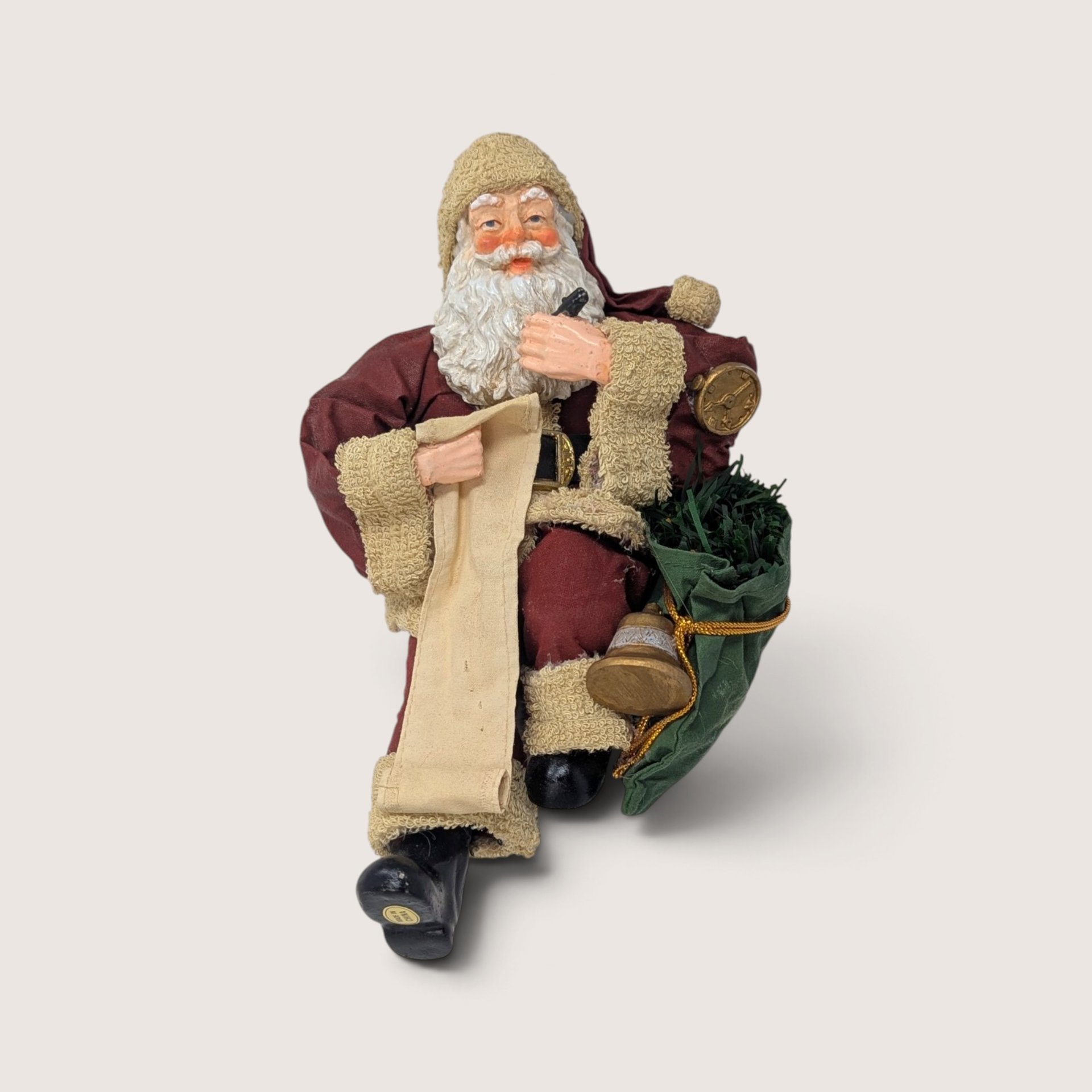 Vintage holiday figurine of Santa Claus sitting down, checking a long list with a pen and holding a toy bag.