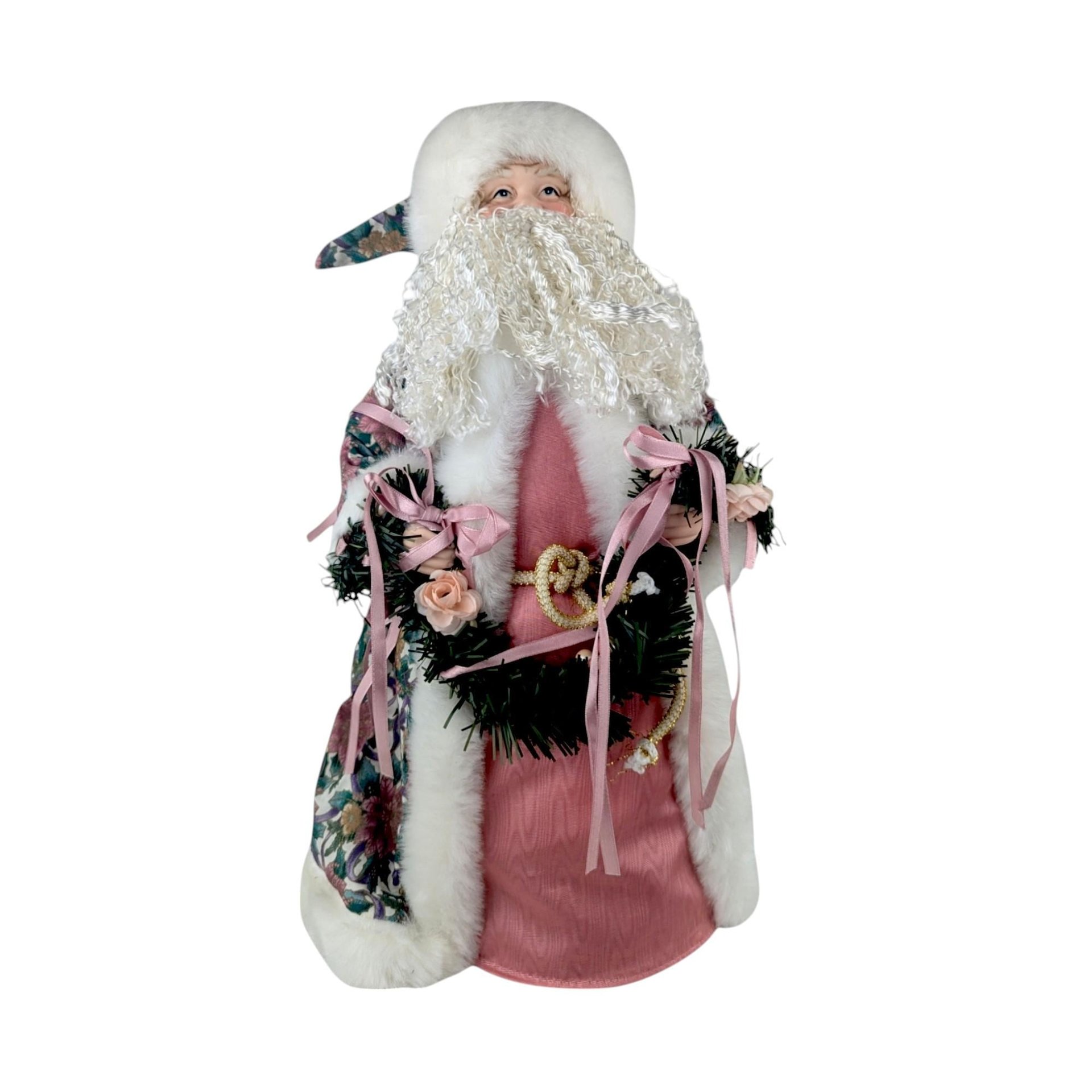 A vertical view of a 16-inch Victorian-style Santa Claus tree topper. He features a long, wavy white beard and wears a soft pink gown with a matching pink floral-patterned overcoat trimmed in white faux fur. He is holding a green tinsel wreath decorated with pink roses and long pink ribbons.