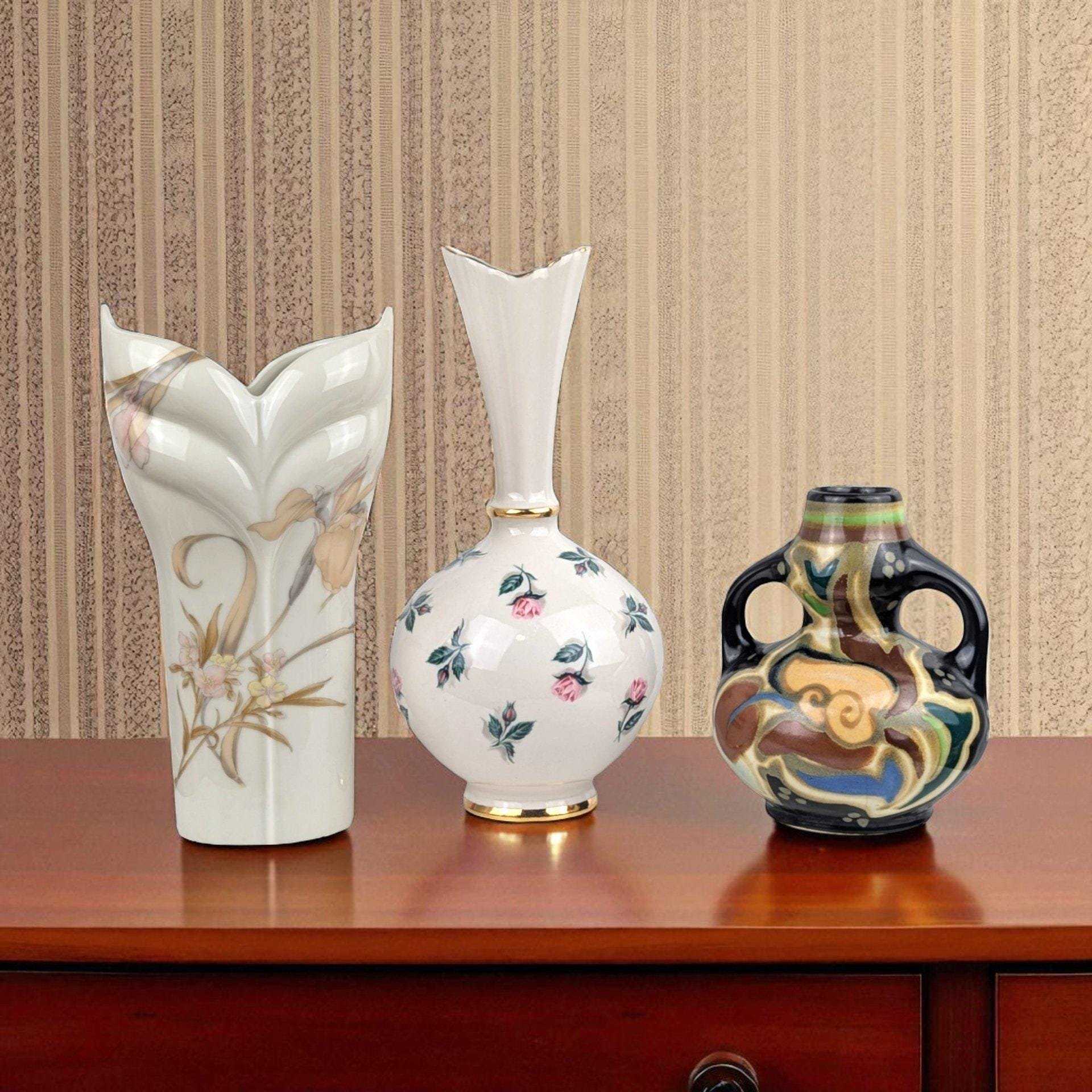 A set of three unique vintage ceramic vases including a tall white vase with pink roses, a white sculptural vase with an orchid design, and a small rounded black vase with colorful abstract patterns.