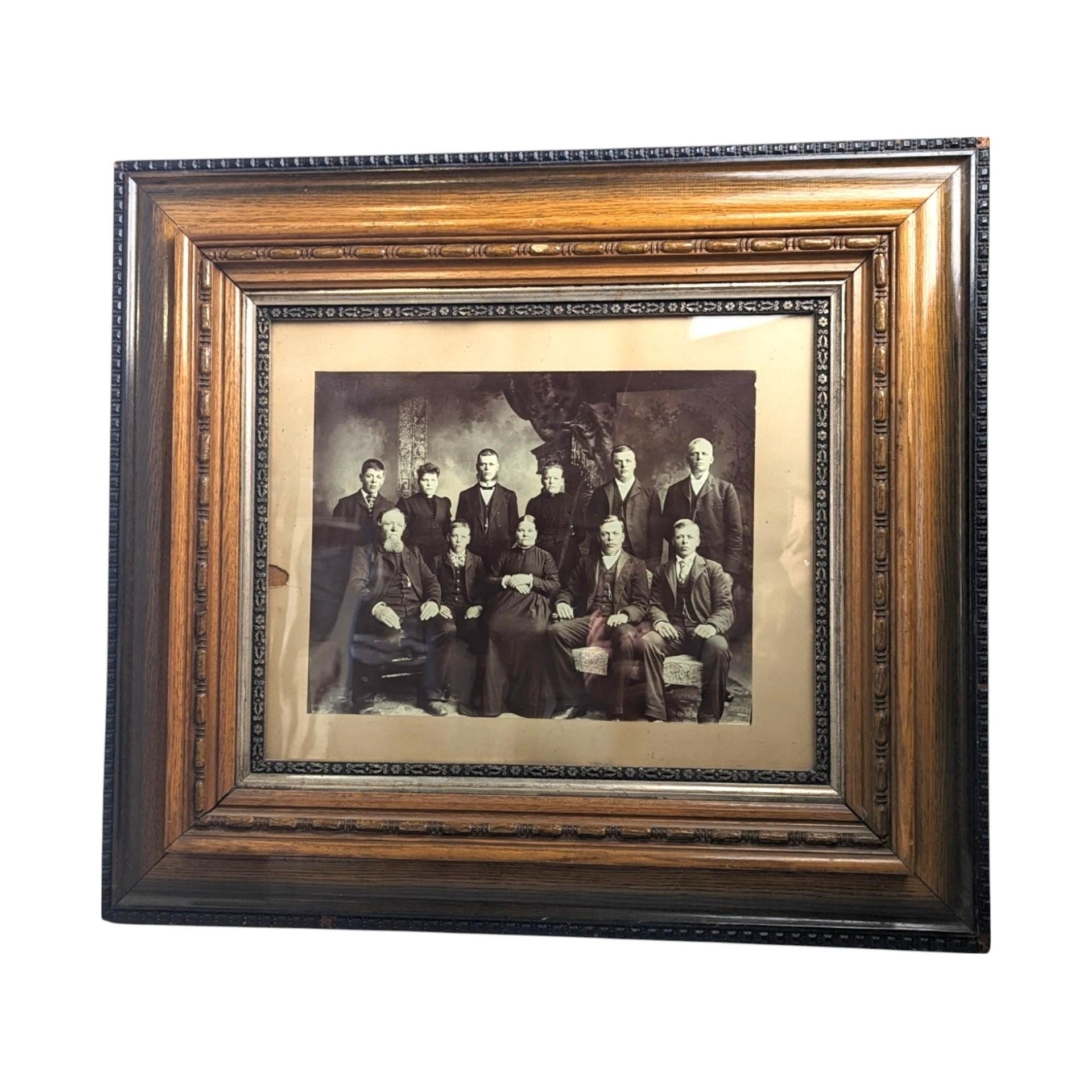 A large, ornate antique wood and gesso picture frame with a gold-toned finish and deep molding, housing a sepia-toned historical photograph of people in formal Victorian attire.