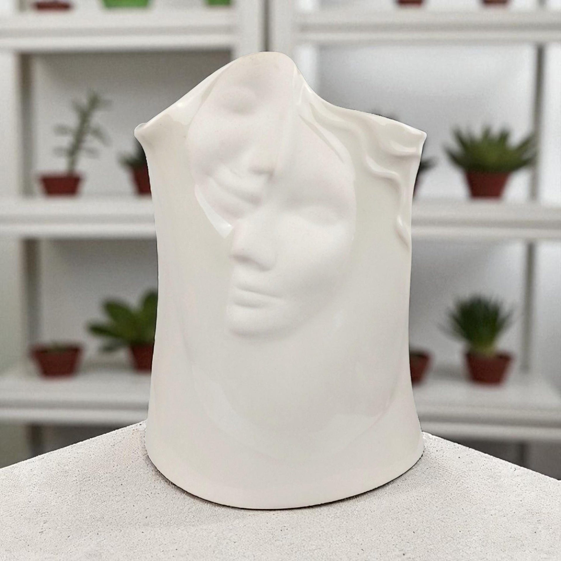 A minimalist, all-white porcelain vase sculpted in the shape of a serene female face. The vase features an elongated neck and a stylized head, with flowing hair textured into the ceramic. The entire piece is finished in a clean, high-gloss white glaze, emphasizing the smooth contours and delicate facial features without the use of color.