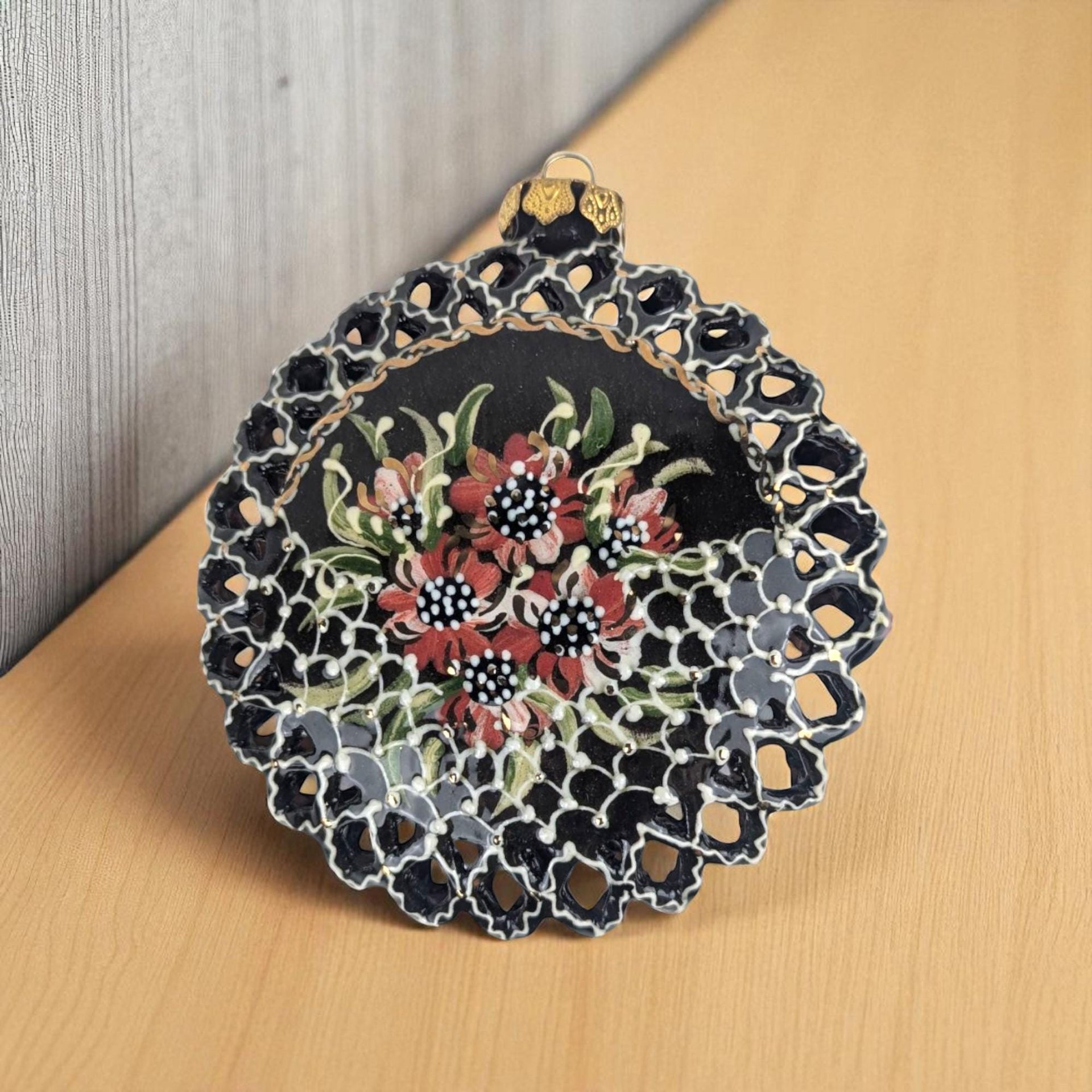A 4-inch round black clay ornament with a delicate reticulated or pierced lattice edge, featuring hand-painted red and white flowers with green leaves.