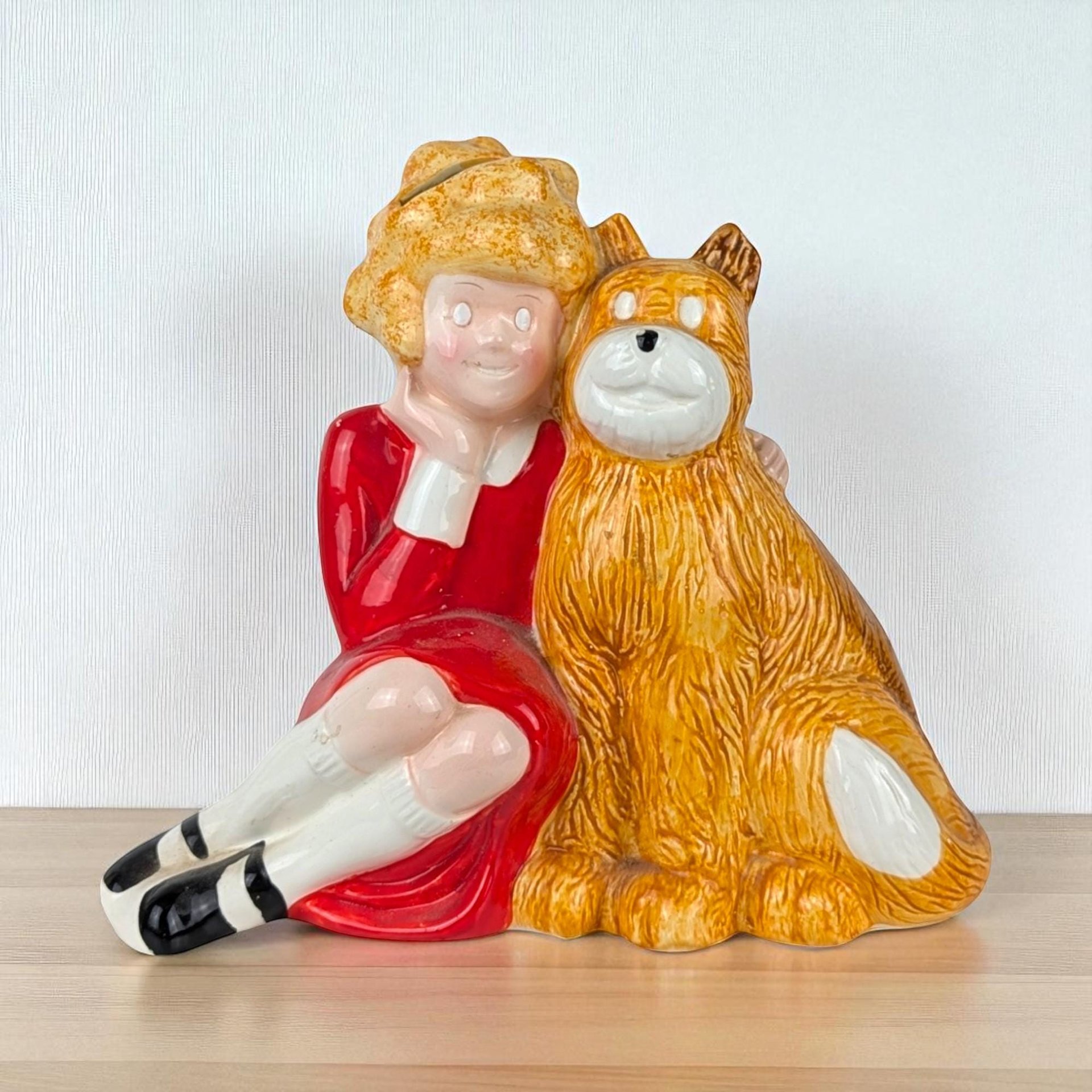 A glossy ceramic coin bank featuring Little Orphan Annie in her signature red dress sitting beside Sandy the dog. The piece displays a smooth, reflective glaze over molded details, capturing the classic character silhouettes in warm red and golden-brown tones.