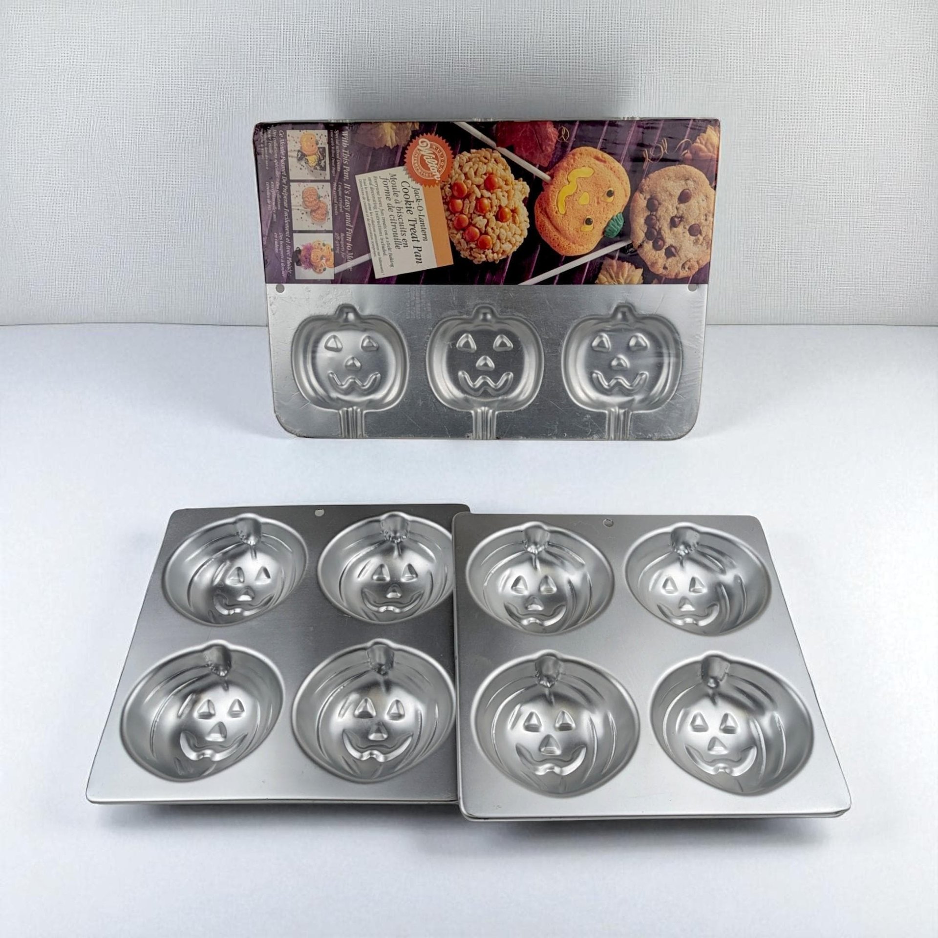 A lot of three silver aluminum baking pans featuring jack-o'-lantern face molds. One pan is a specialized cookie treat pan with stick slots and original cardboard packaging; the other two are matching four-cavity muffin or mini-cake pans.