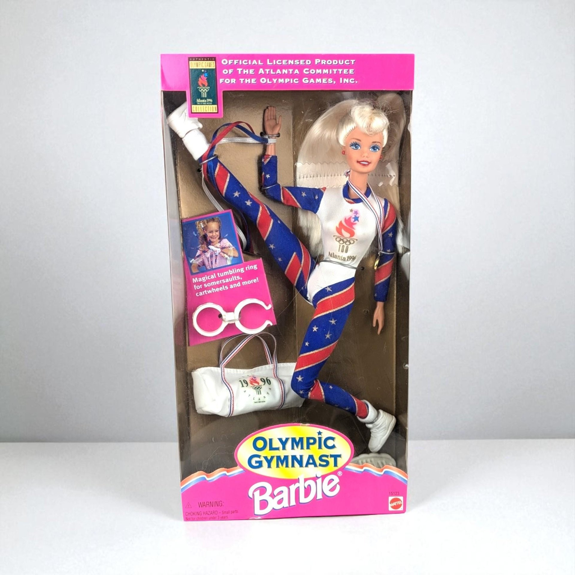 The Olympic Gymnast Barbie is pictured in her original packaging, posed in a dynamic split. She wears a patriotic red, white, and blue leotard with star accents and carries a white gym bag. A pink tumbling ring and gold medal accessory are visible within the box.