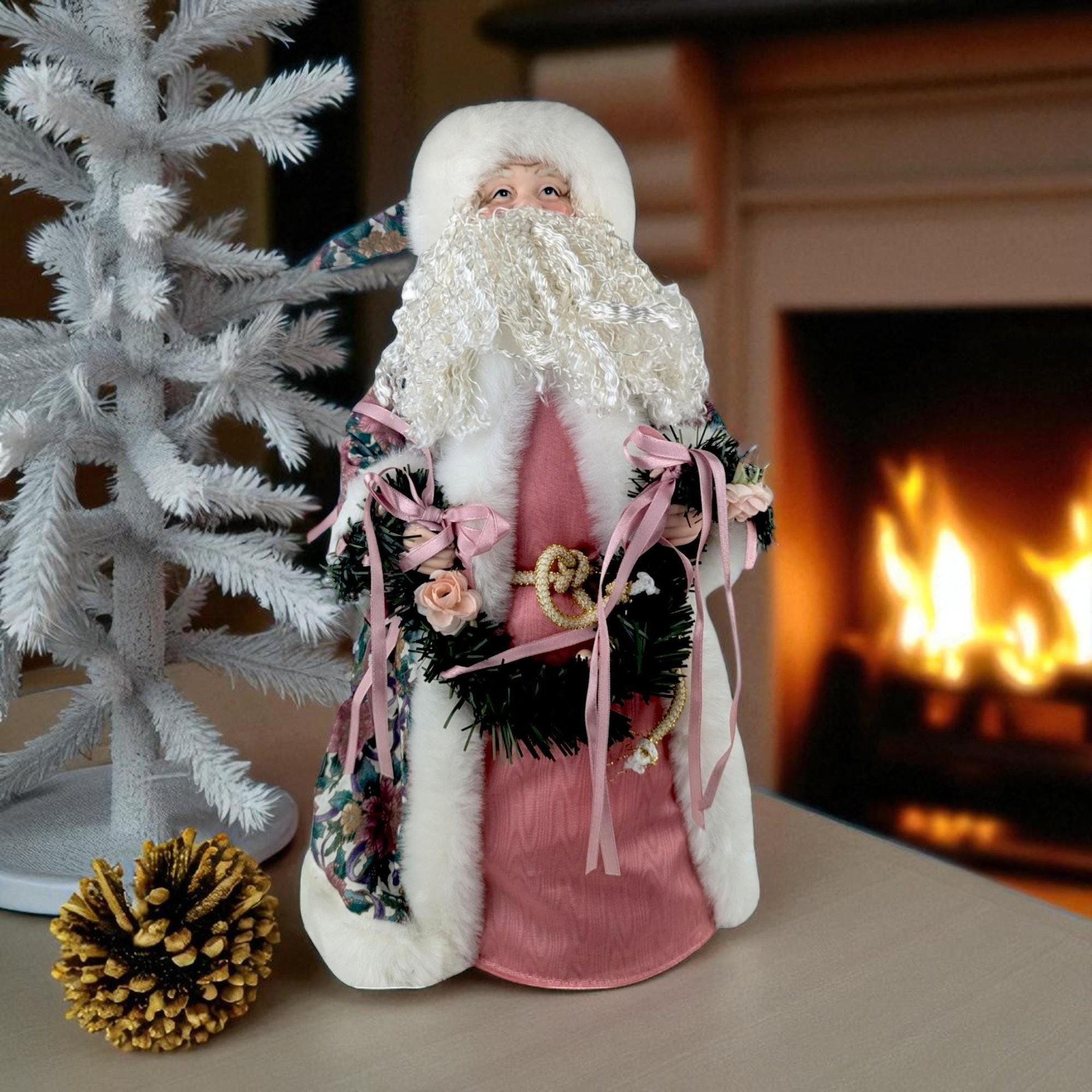 Intricate Christmas tree topper featuring a Santa figure in a Victorian-style pale pink robe. The robe is adorned with delicate floral patterns and trimmed with soft white faux fur. He has a realistic, hand-painted porcelain face and carries a small bundle of frosted greenery. The silhouette is conical and elegant.