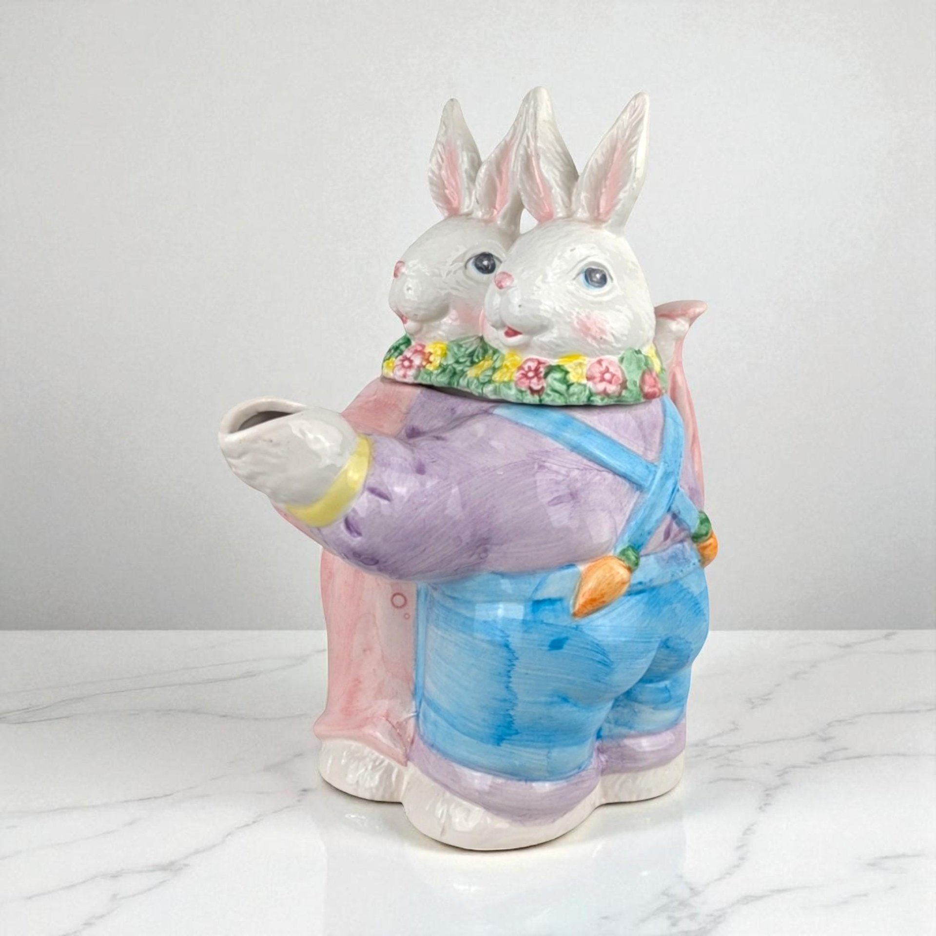 Ceramic teapot featuring a group of three white rabbits in blue and yellow clothing. The rabbits form a circular, dancing silhouette. Glossy glaze texture with a curved handle and spout. Pastel color palette of blue, yellow, and cream.