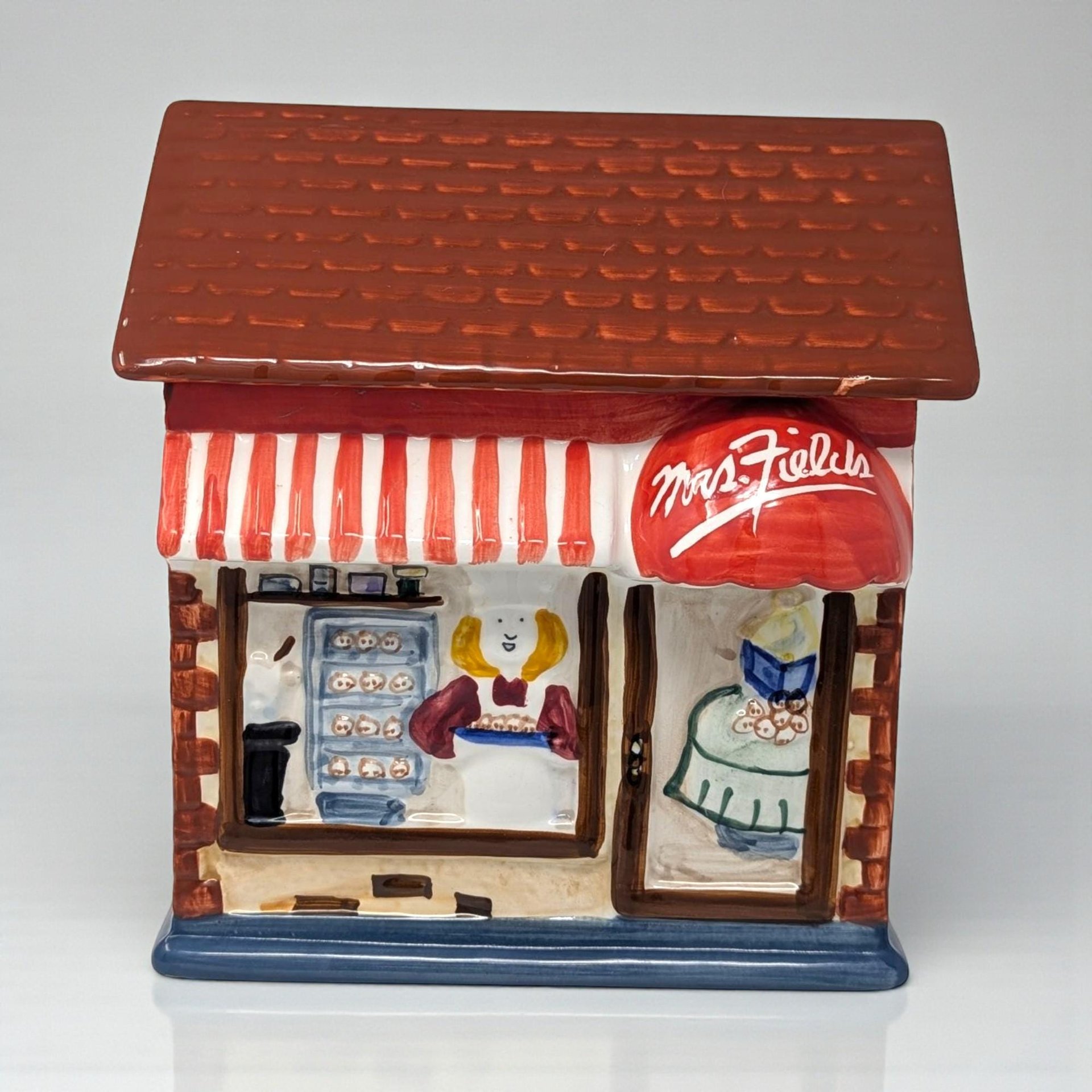 A ceramic Mrs. Fields cookie shop cookie jar. The jar is shaped like a storefront with a brown shingled roof, a red and white striped awning, and a red dome awning with the Mrs. Fields logo. The front depicts a baker inside among trays of cookies.