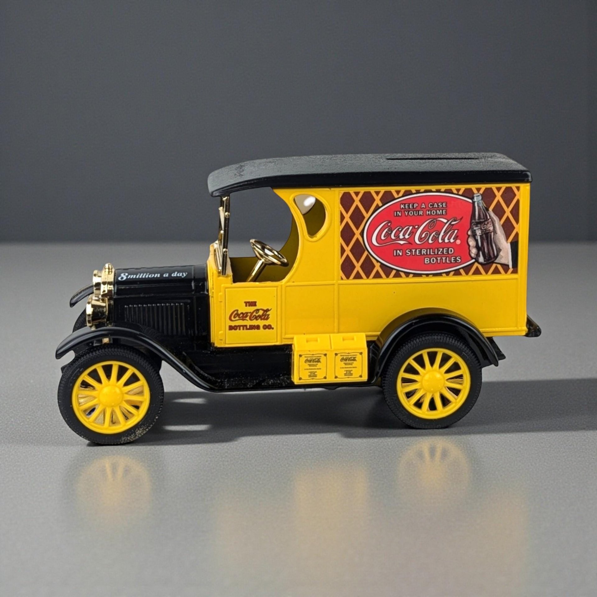 A die-cast metal bank in the shape of a vintage yellow and black Coca-Cola delivery truck. The vehicle features yellow spoked wheels, gold-toned hardware, and detailed side graphics including a red Coca-Cola logo and a "8 million a day" slogan on the hood.