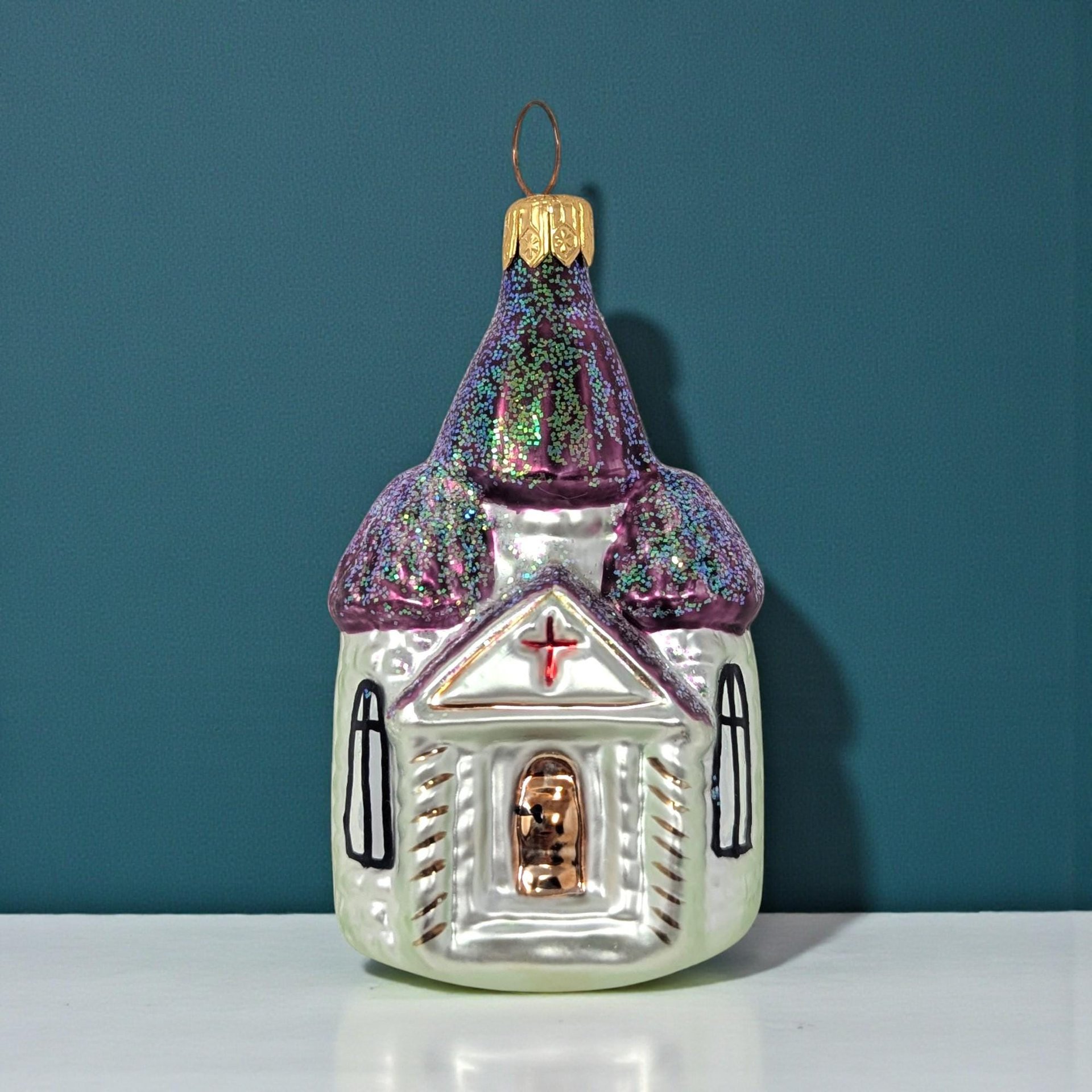 A hand-painted glass church ornament with a pearlescent white body and shimmering purple domes. The spires are dusted with iridescent green glitter, featuring a gold-toned topper and a small red cross above the entryway.
