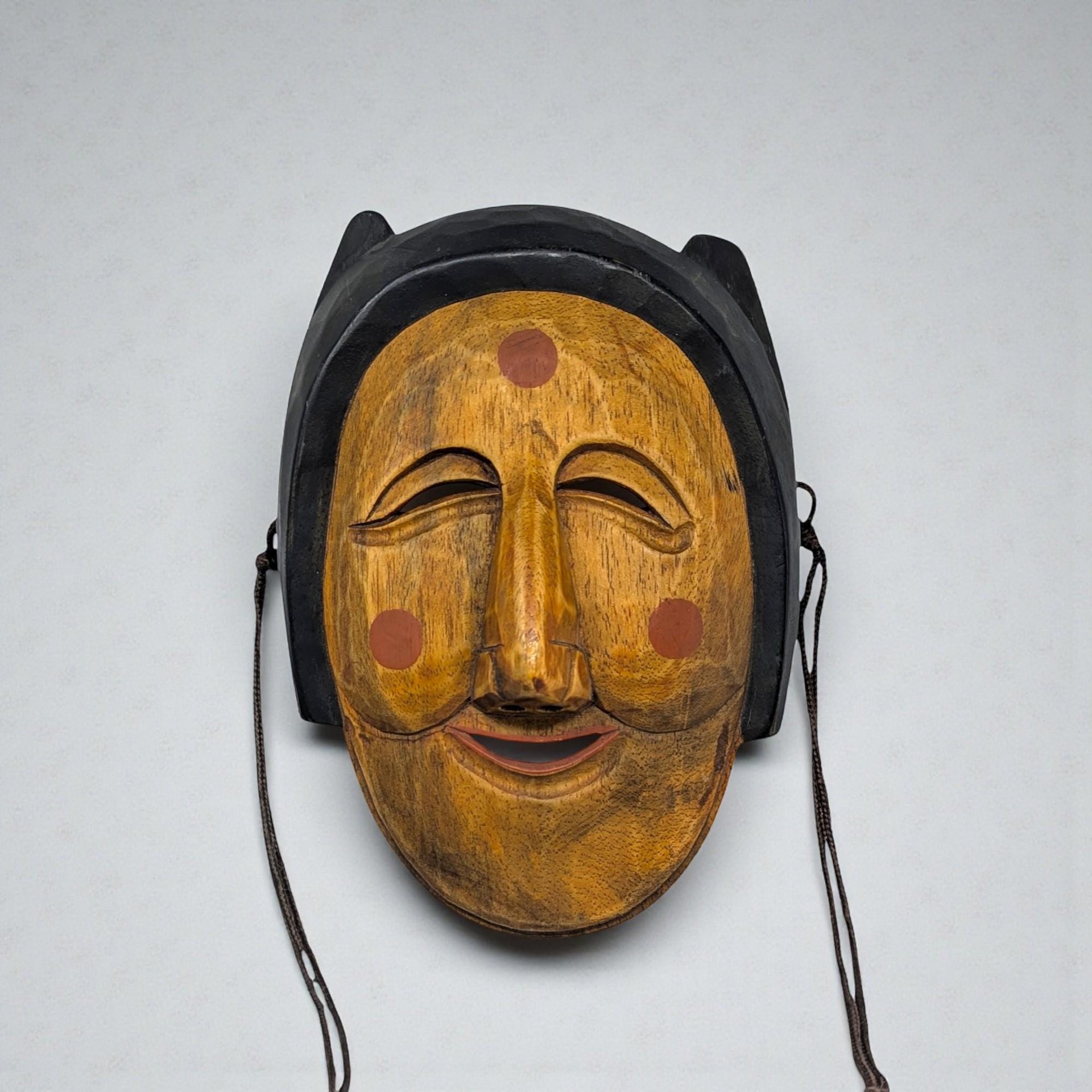 A hand-carved wooden mask with a deep mahogany finish. The face features elongated features, slit eyes, and a prominent nose, with subtle geometric patterns etched along the forehead and cheeks.