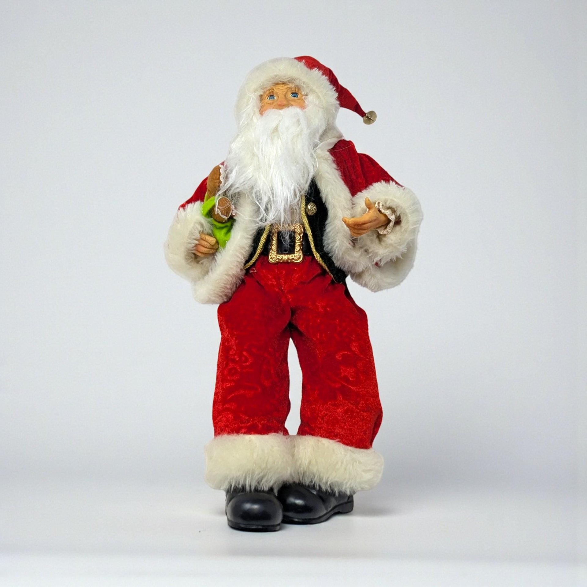 A seated Santa figure in an elf-style pose, wearing a red velvet suit with green trim and a tall pointed hat. He holds a stuffed gingerbread man and features long, dangling legs with pointed black boots.