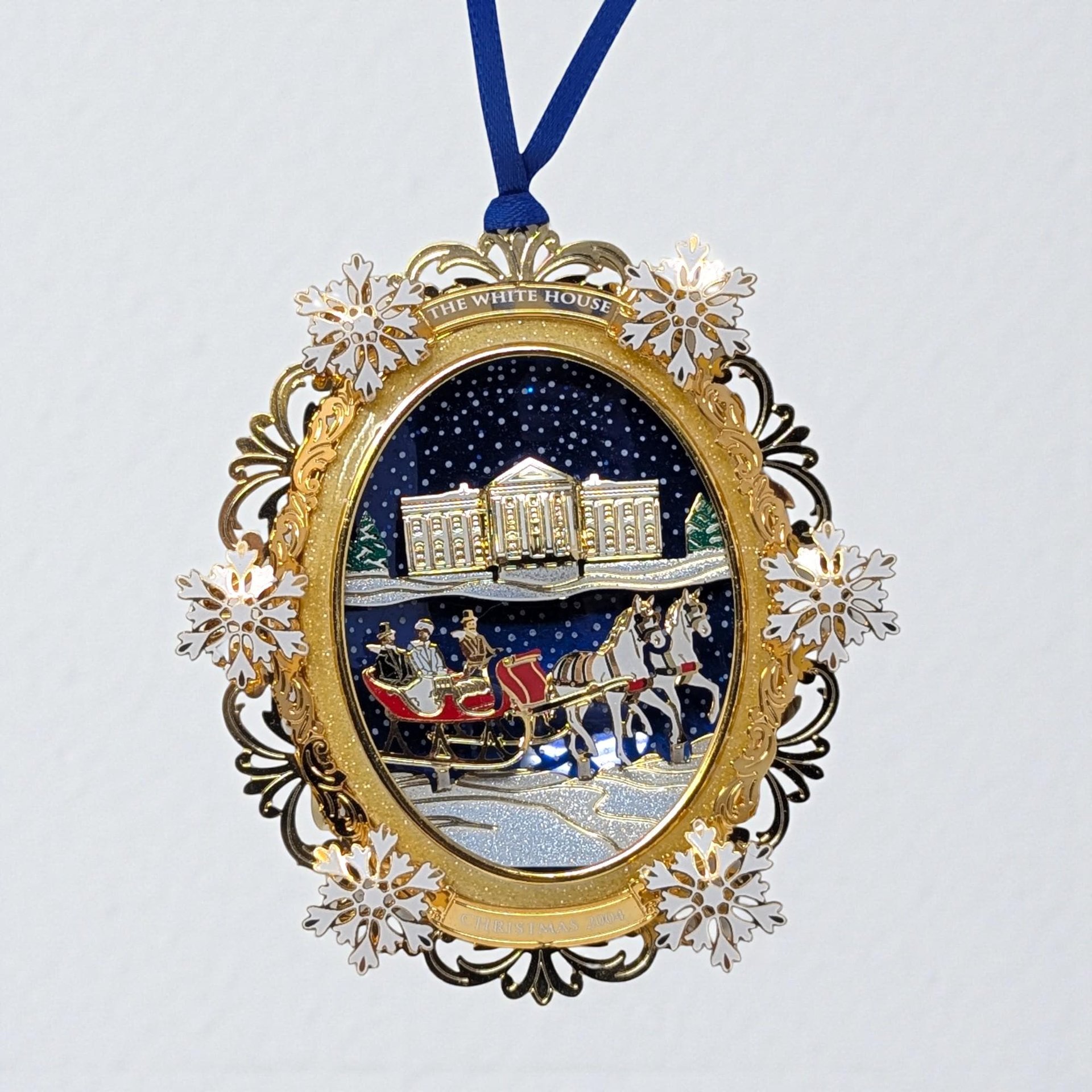 A detailed gold-tone metal ornament depicting a snowy winter sleigh ride. A horse-drawn carriage carries a couple through an intricate archway of frosted trees and architectural elements, suspended by a blue ribbon.