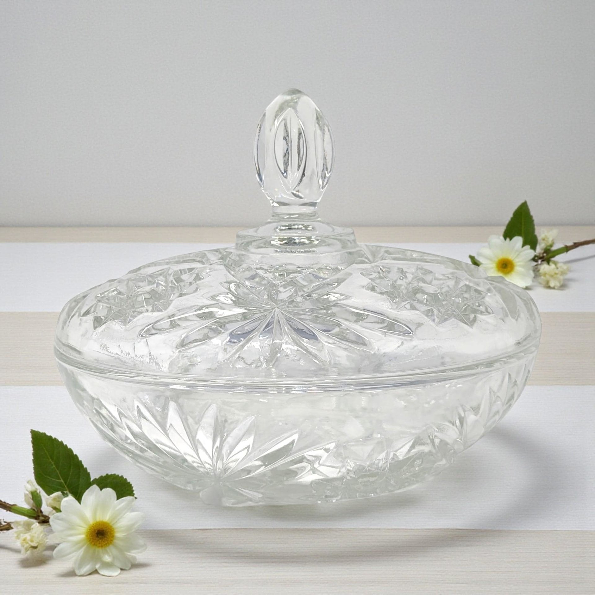 A clear glass serving dish with a matching lid featuring a starburst and pineapple pattern. The bowl has a scalloped rim and a pedestal-like base, while the lid is topped with a faceted pointed finial.