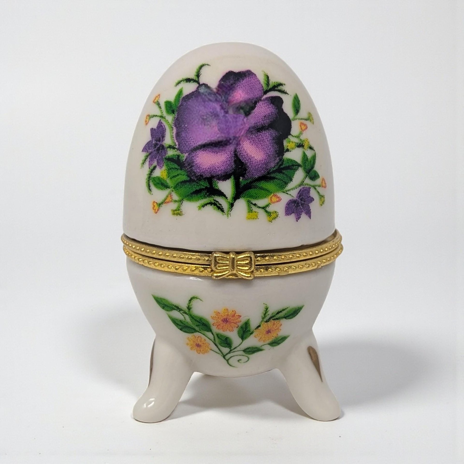 A small, egg-shaped white ceramic box with a hinged gold metal closure. The exterior is decorated with delicate purple violet sprays and green leaves on both the lid and base.