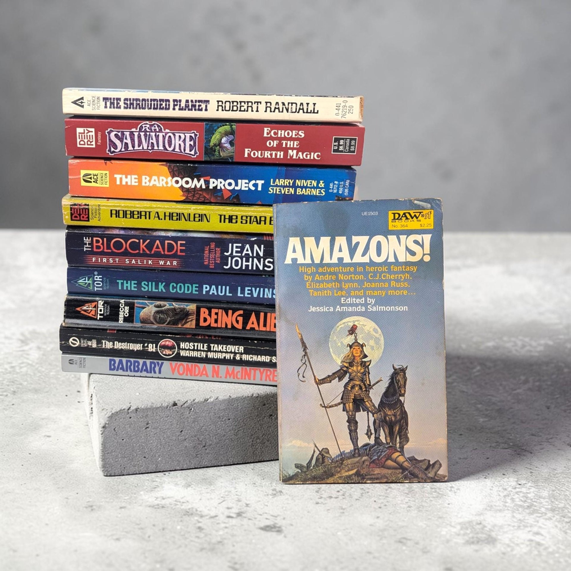 A stack of nine vintage science fiction and fantasy paperbacks with multi-colored spines. The book on top, "Amazons!" edited by Jessica Amanda Salmonson, is also leaned against the front of the stack, showcasing cover art of a woman in golden armor standing under a full moon.