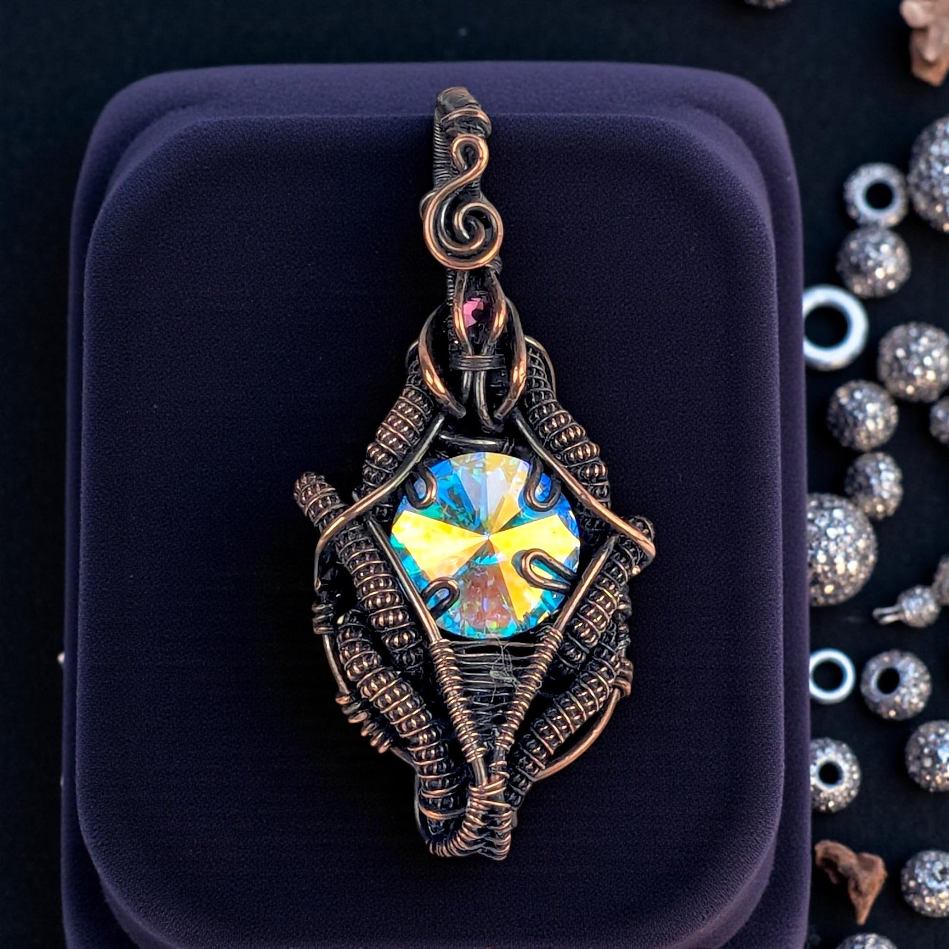 A diamond-shaped pendant featuring a large, iridescent rivoli crystal that shifts between gold, blue, and teal. It is encased in dense, antiqued copper wire weaving with a small purple crystal accent and a scrolled bail.