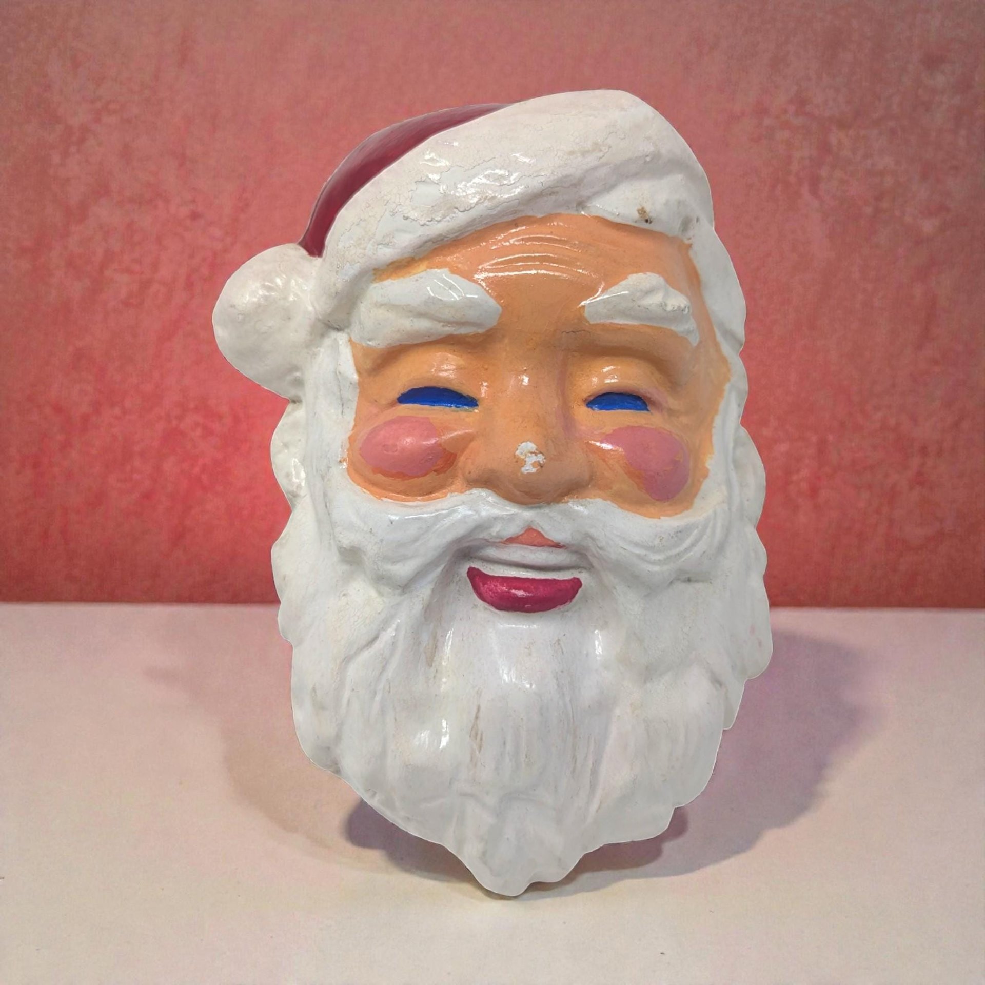 Chalkware Santa head wall plaque with a textured white beard and a bright red hat. He features a traditional hand-painted face and mall metal loops ties with string on the back for hanging.