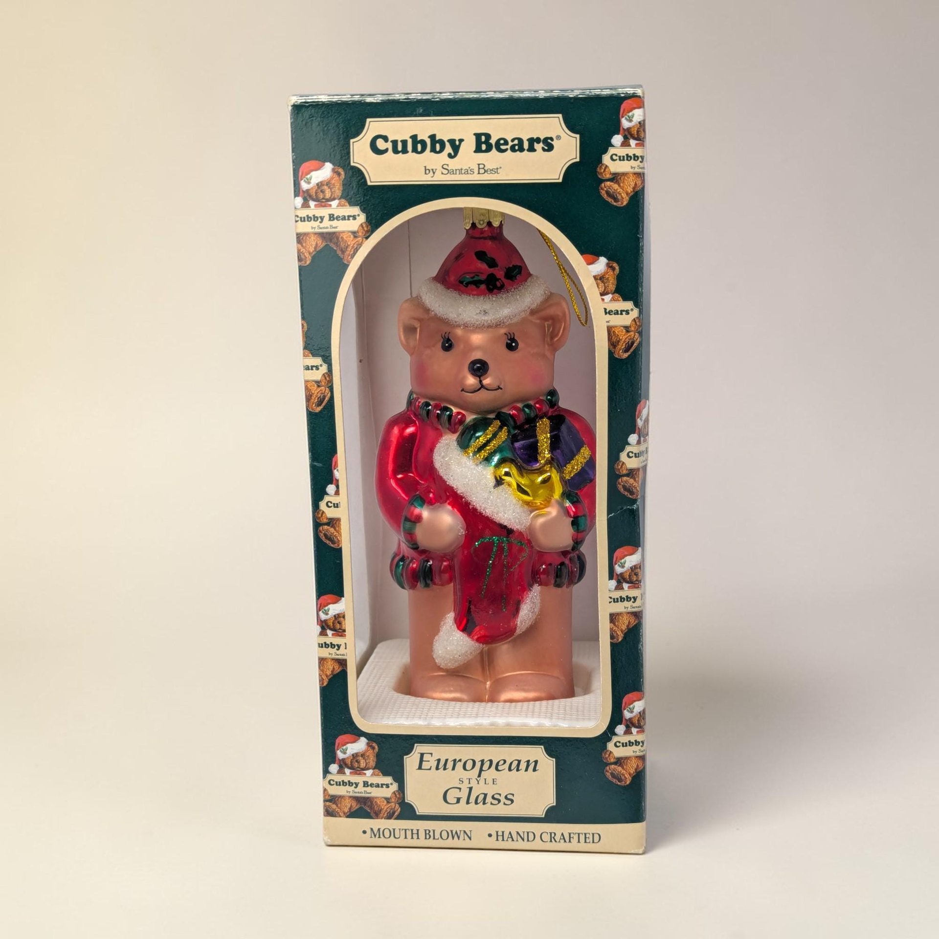 A large glass bear ornament wearing a red and white Santa hat and holding a green present. The texture is a shimmering, metallic hand-painted glass with a tall, sculpted silhouette and a gold hanging loop.