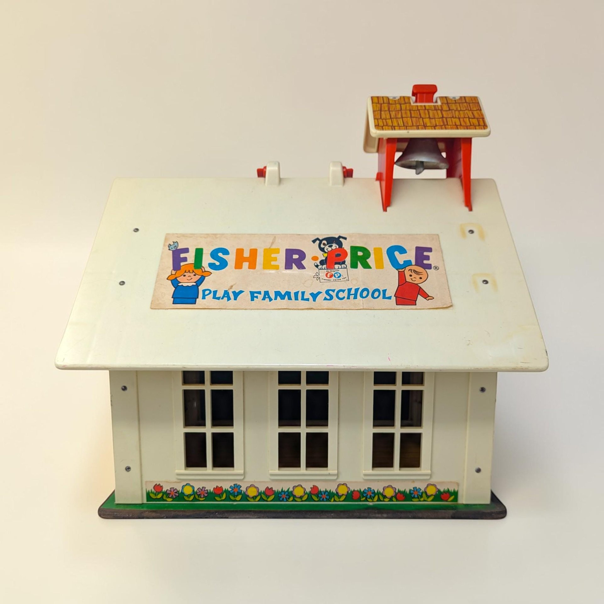 A cream-roofed toy schoolhouse with a red base and carry handle. The texture is a mix of sturdy molded plastic and lithographed paper siding in a rectangular, boxy silhouette.