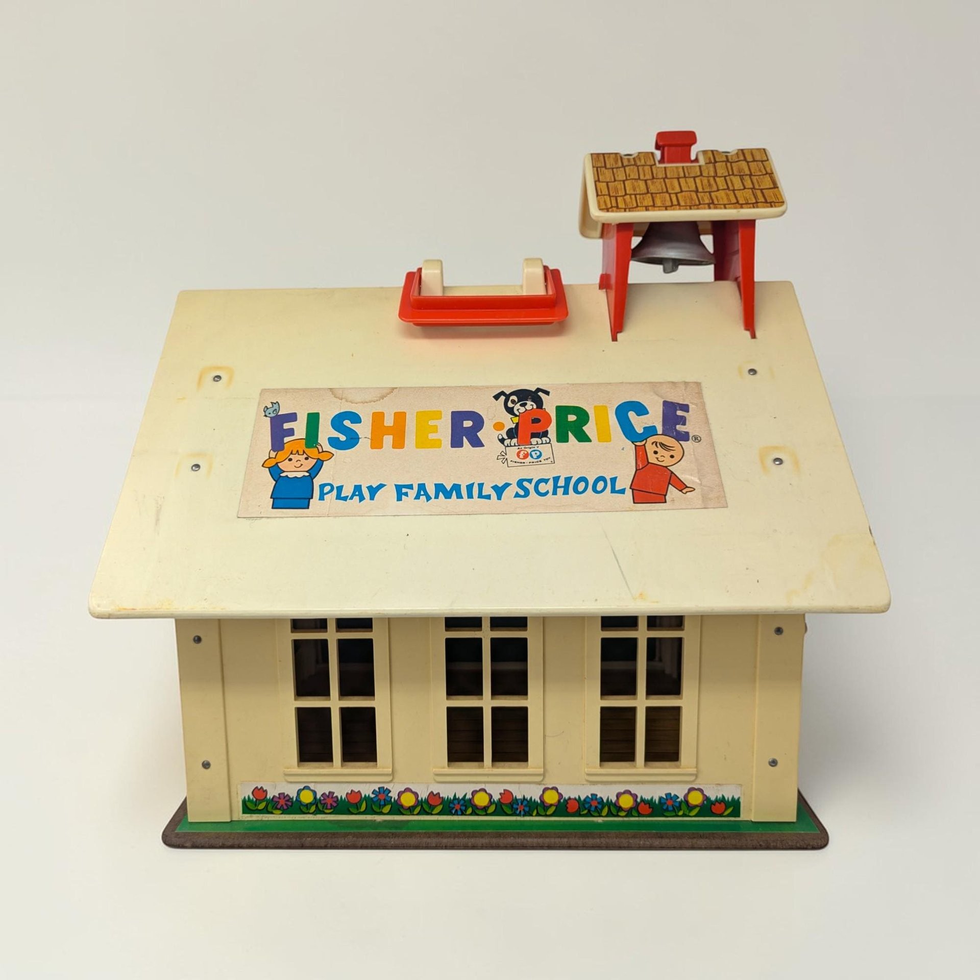 A cream-roofed toy schoolhouse with a red base and carry handle. The texture is a mix of sturdy molded plastic and lithographed paper siding in a rectangular, boxy silhouette.