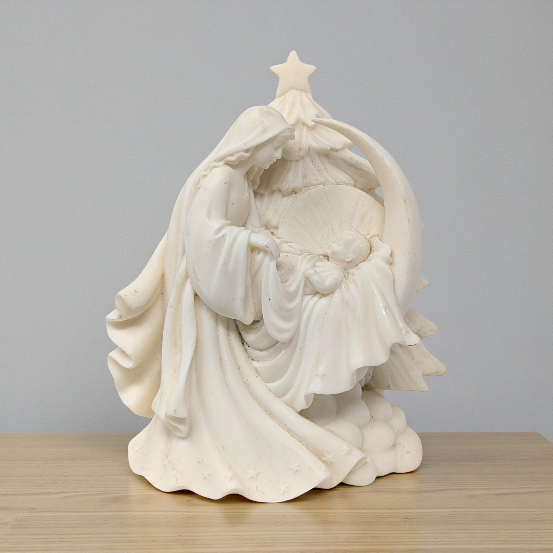 A cream-colored resin figurine of the Holy Family. Mary and Joseph are depicted in a tender, protective pose over the infant Jesus, framed by a smooth, contemporary stylized arch with a clean matte finish.