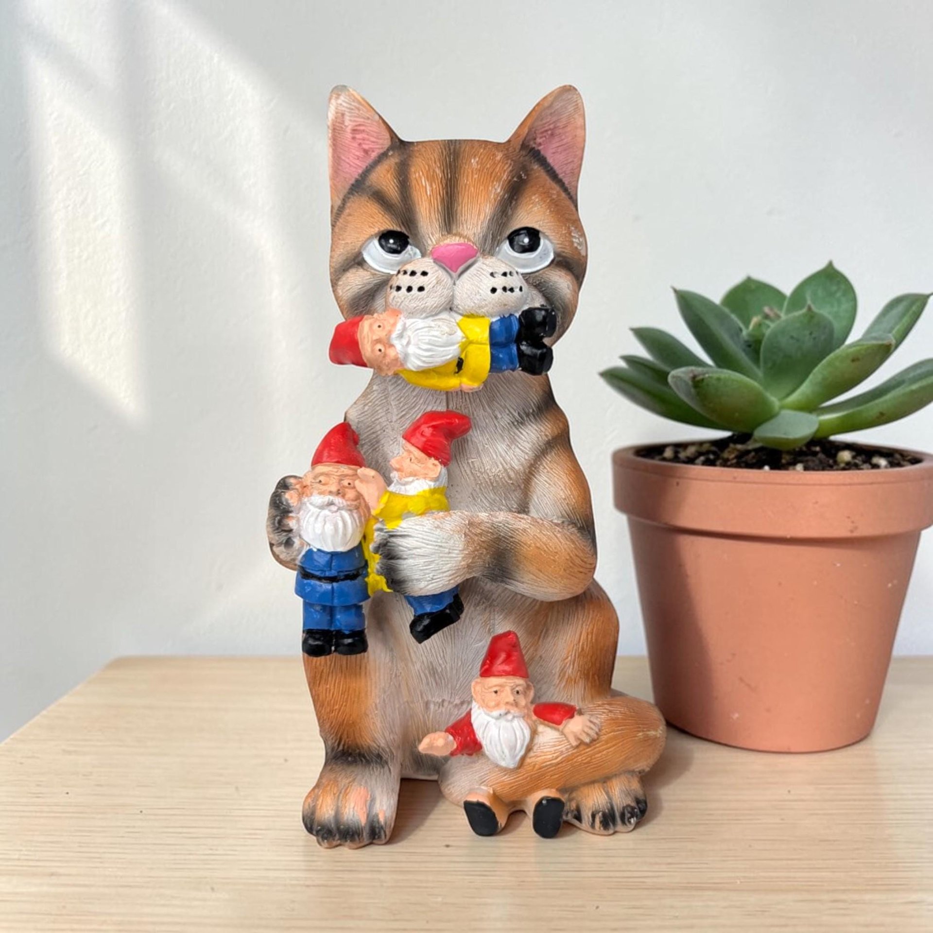 A textured resin figurine of a brown tabby cat with a pink nose, sitting upright while holding and biting several small garden gnomes with red hats. The cat has a mischievous expression and a sculpted, fur-like finish.