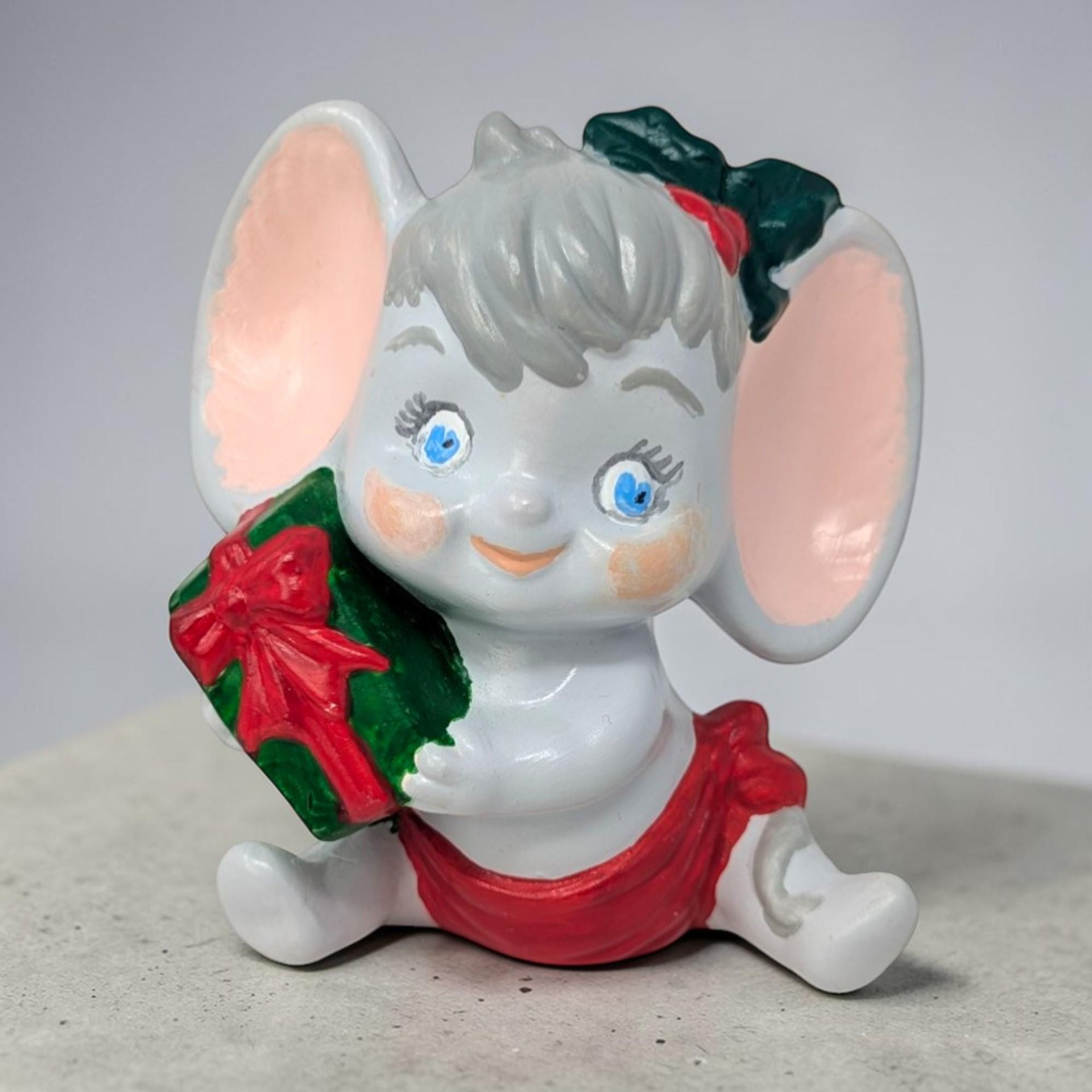 A hand-painted ceramic figurine of a gray mouse with large rounded ears, wearing a red Santa hat and a green scarf. The mouse is sitting and holding a small yellow candle, featuring a glossy glazed finish and a sweet, wide-eyed expression.
