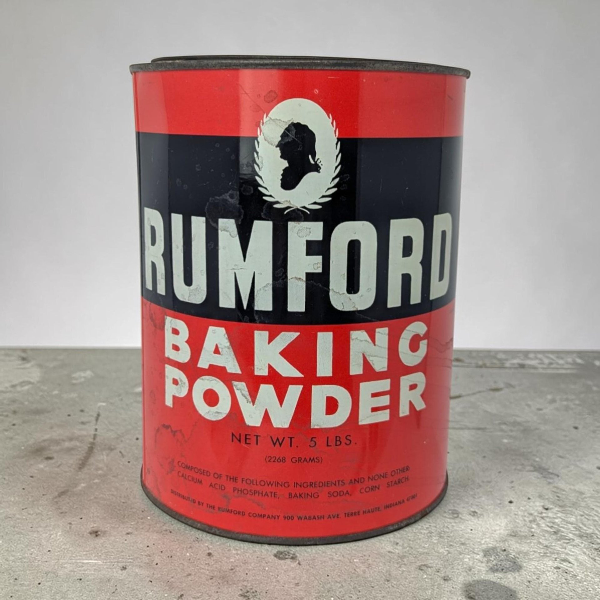 A large cylindrical metal tin with a bold red and black label. White block lettering reads RUMFORD BAKING POWDER. A white oval at the top contains a black silhouette of a man's profile framed by a laurel wreath.