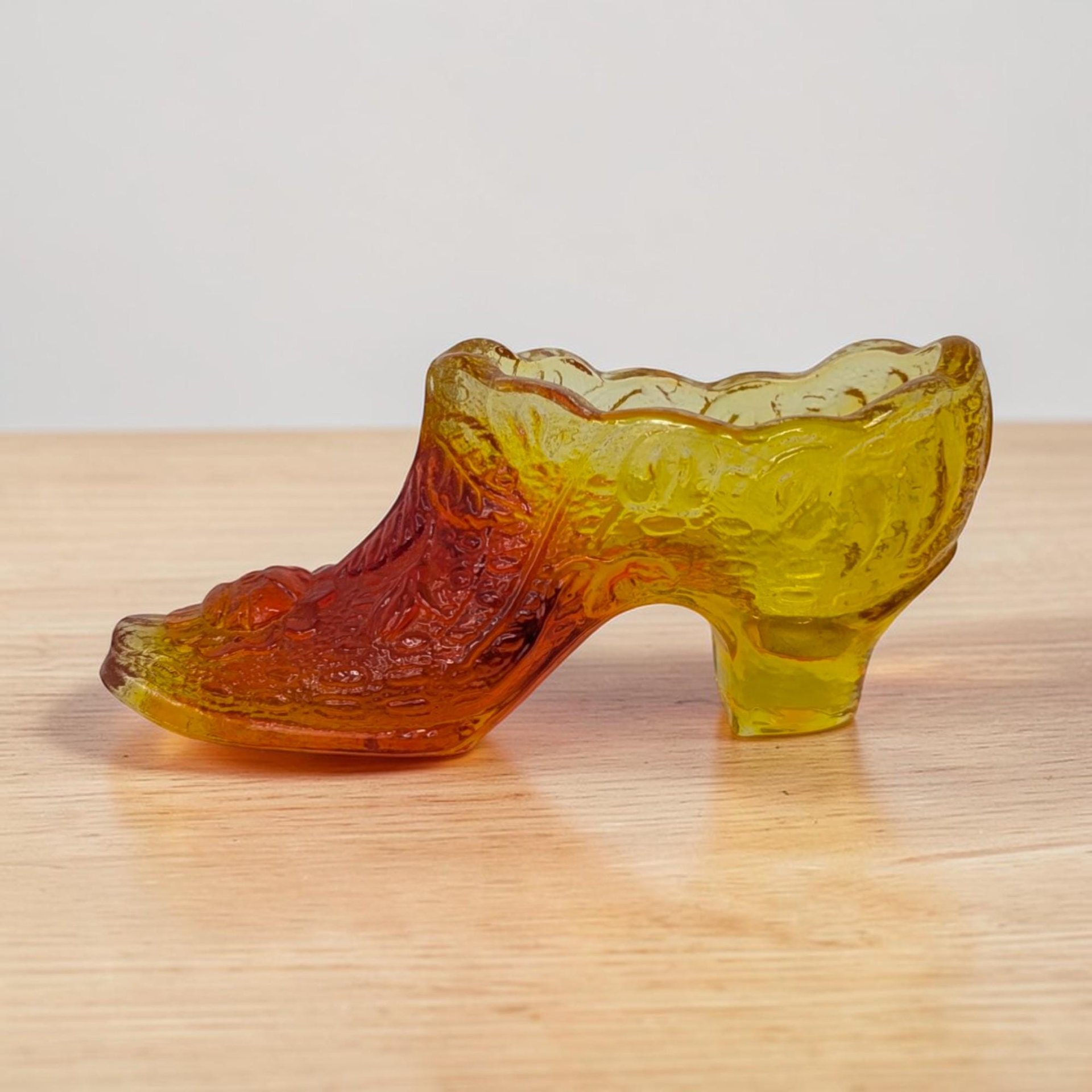 A heavy glass slipper with a ruffled rim and pointed toe. The color transitions from a deep, fiery red at the top to a golden honey amber at the base. The surface features a decorative embossed pattern.
