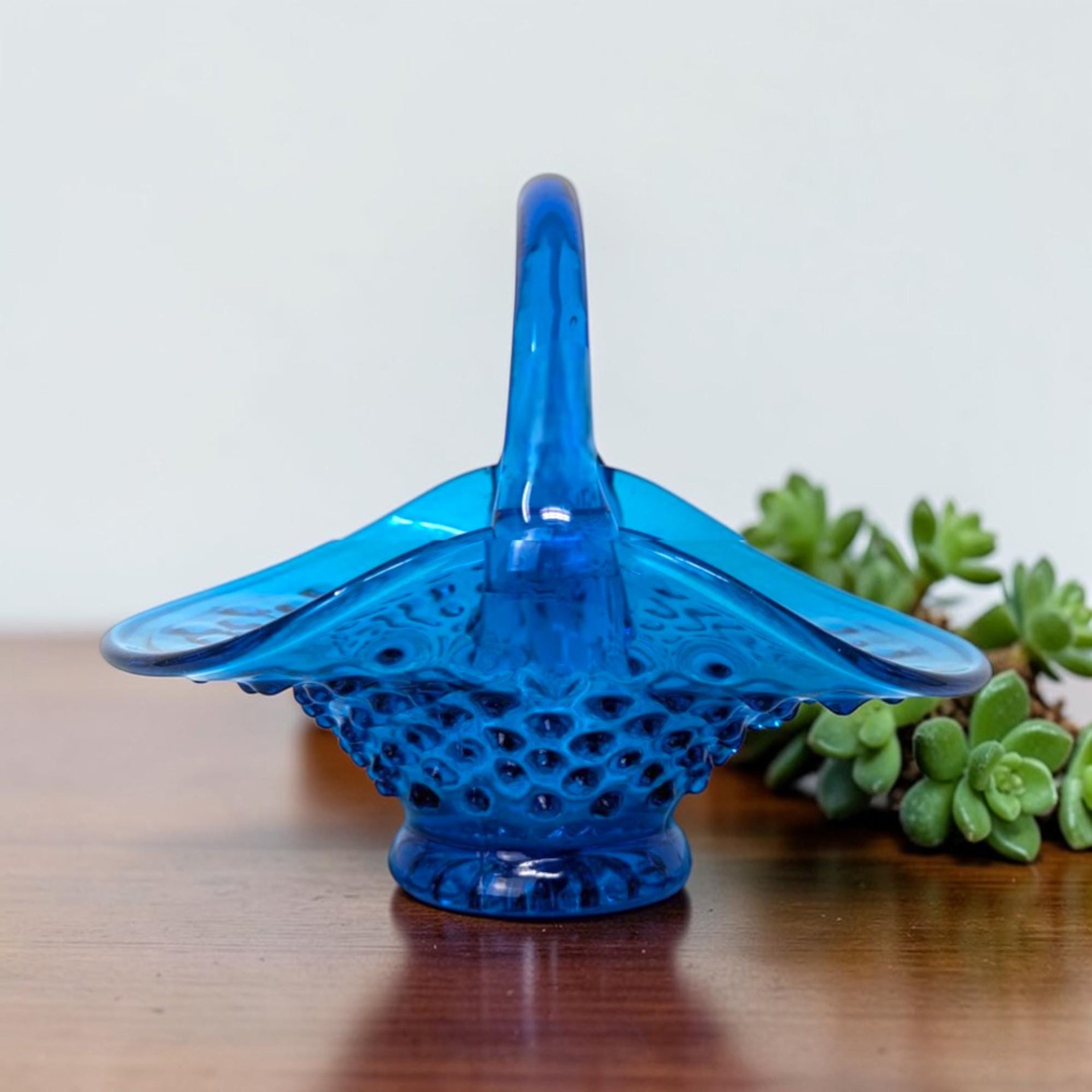 Translucent cerulean blue glass basket featuring a dense hobnail texture on the rounded base and a smooth, arched applied handle. The rim is dramatically flared and ruffled into a wide boat shape.