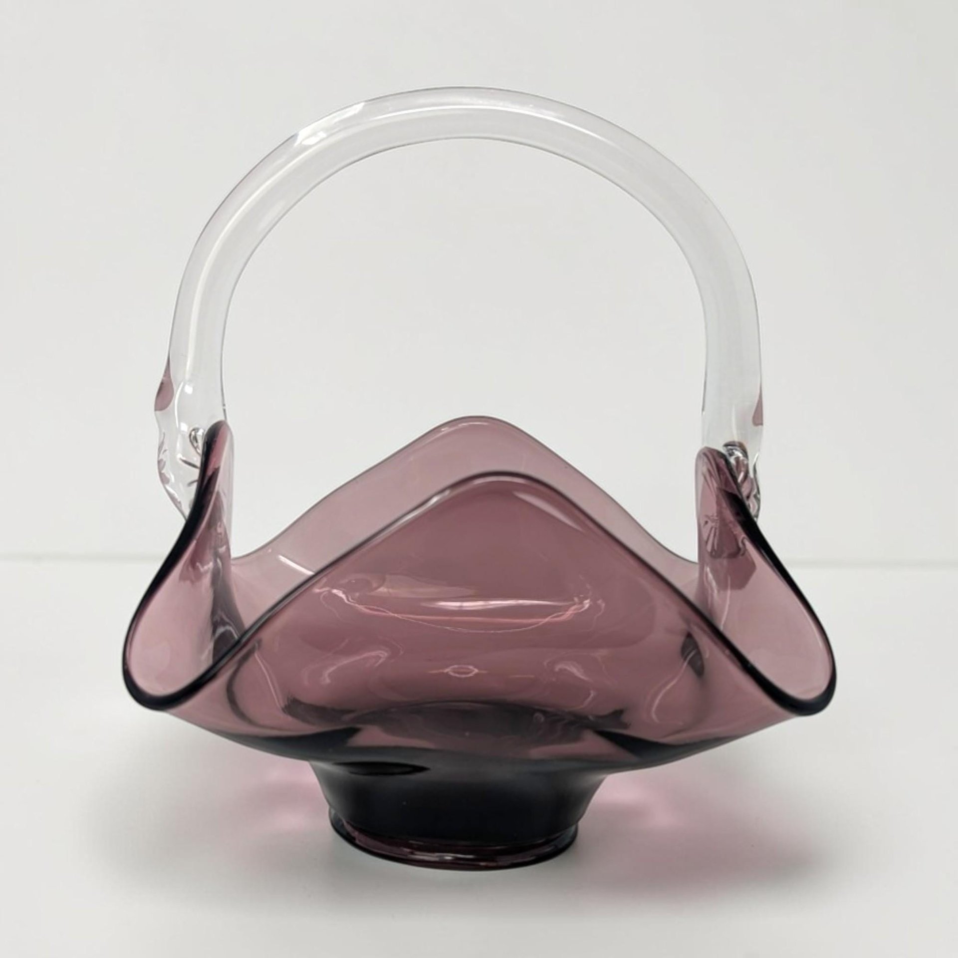 A hand-blown amethyst glass basket with a ruffled handkerchief-style rim and a clear glass arched handle. The deep purple body transitions from dark to translucent, featuring a smooth, polished texture and a pedestal base.