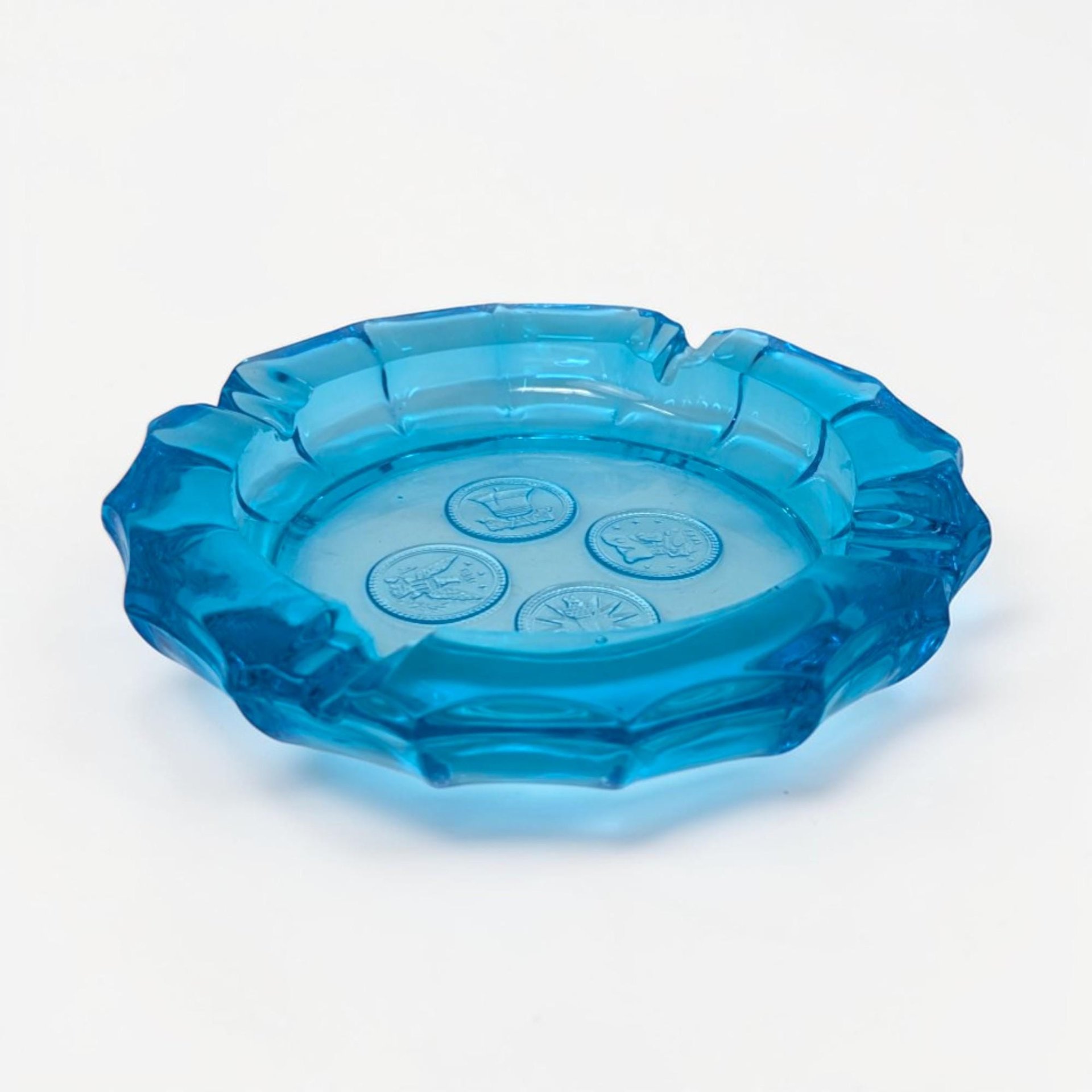 A thick, translucent ice blue glass ashtray with a scalloped edge. Four circular medallions featuring frosted patriotic "coin" motifs, including an eagle and a torch, are embossed into the center of the heavy glass base.