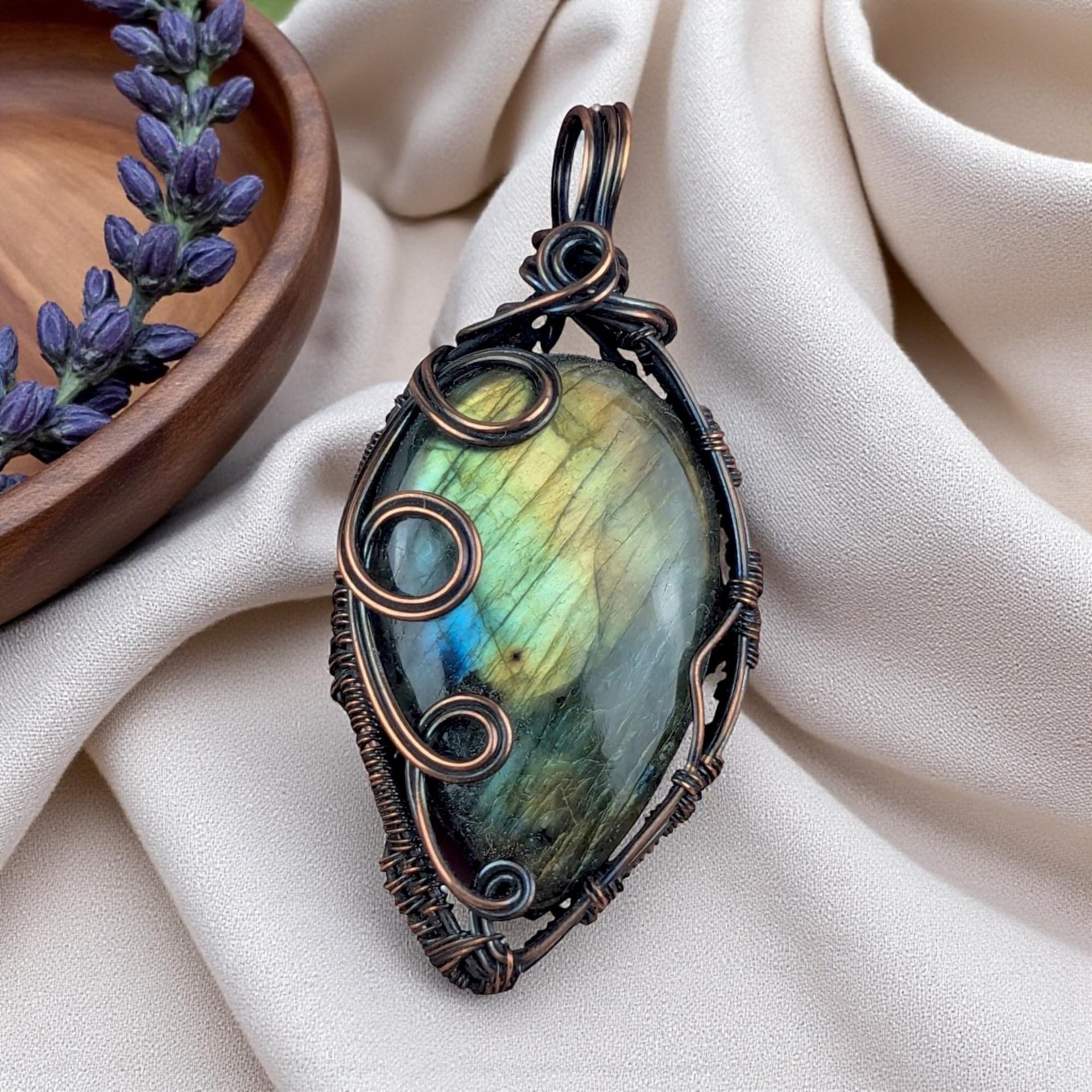 A large teardrop labradorite stone with yellow and green hues. It is wrapped in dark oxidized copper wire featuring three prominent polished spirals on the left side and a woven bail at the top.