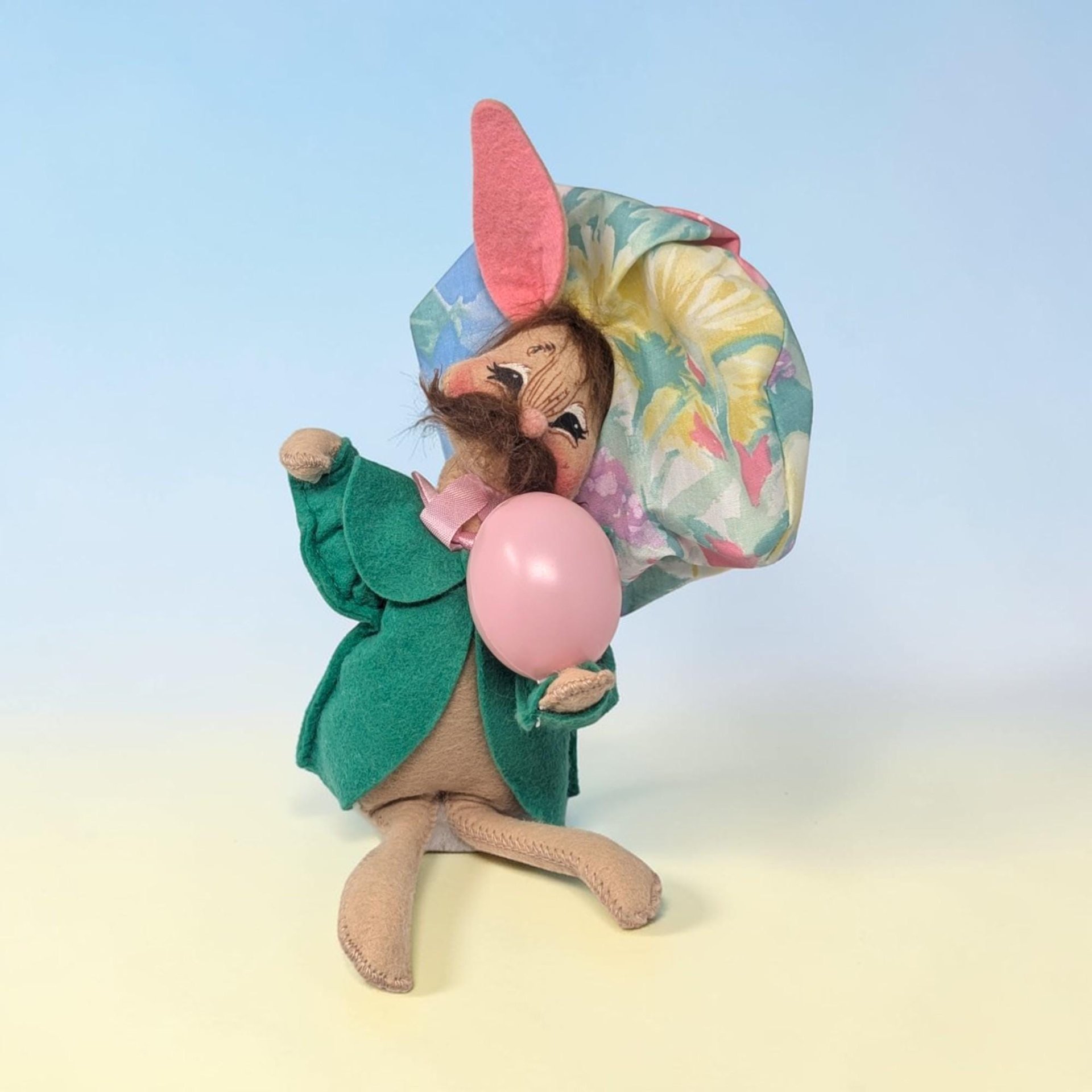 Tan felt bunny wearing a green coat and a large pastel floral beret. He has a dark brown mustache and holds a pink plastic egg. One tall ear is lined in pink felt. He is in a sitting pose against a blue and yellow background.